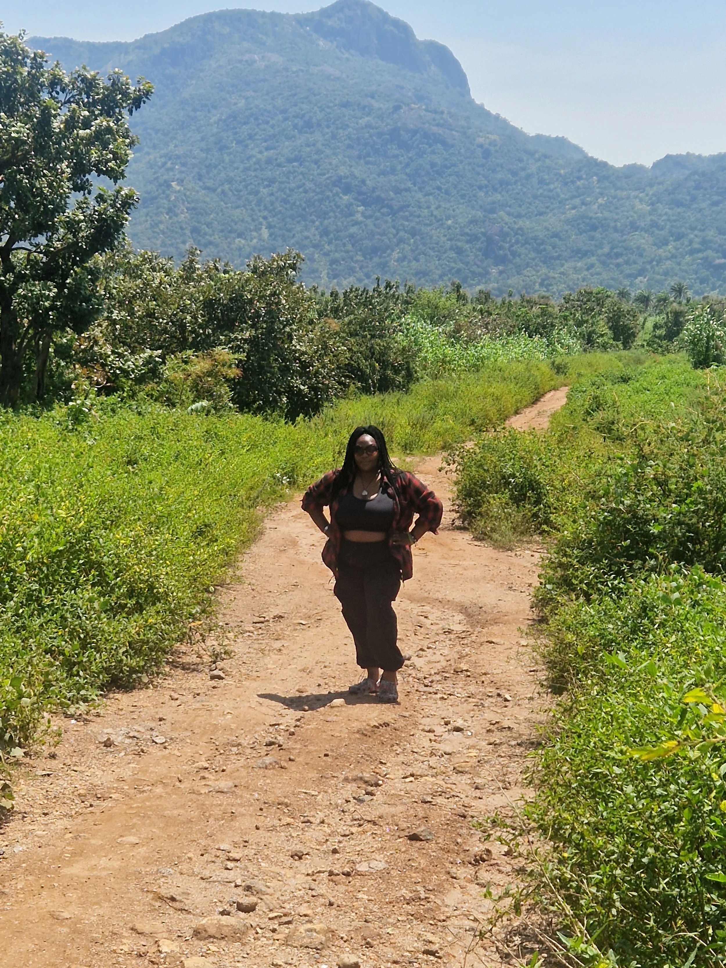 A large mural of a woman with auburn hair, wearing a white sleeveless top, pointing to the right, painted on the side of a tall building. Trees with dark green leaves partially obscure the view.