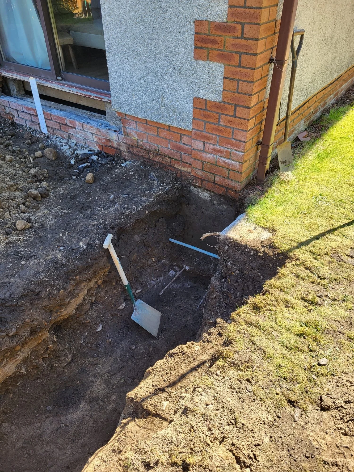 A construction scene showing a dug trench near the corner of a house with brick and stucco exterior walls. Tools like a shovel and a pickaxe are in the trench. The area is outdoors with grass on one side.