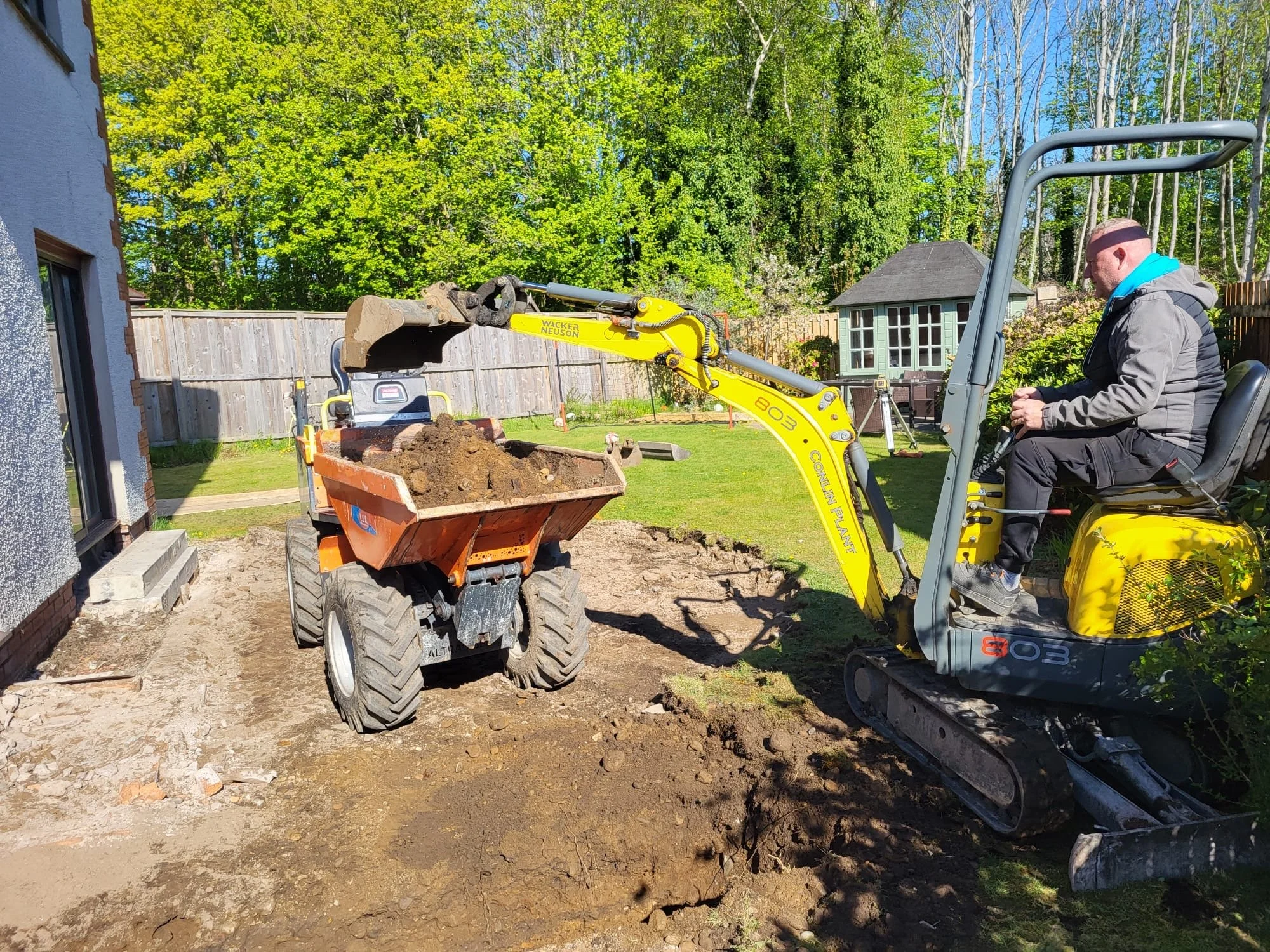 A construction worker operates a small yellow excavator that is digging up earth in a backyard, with a wheelbarrow filled with dirt nearby, and a wooden fence, trees, and a small shed in the background.