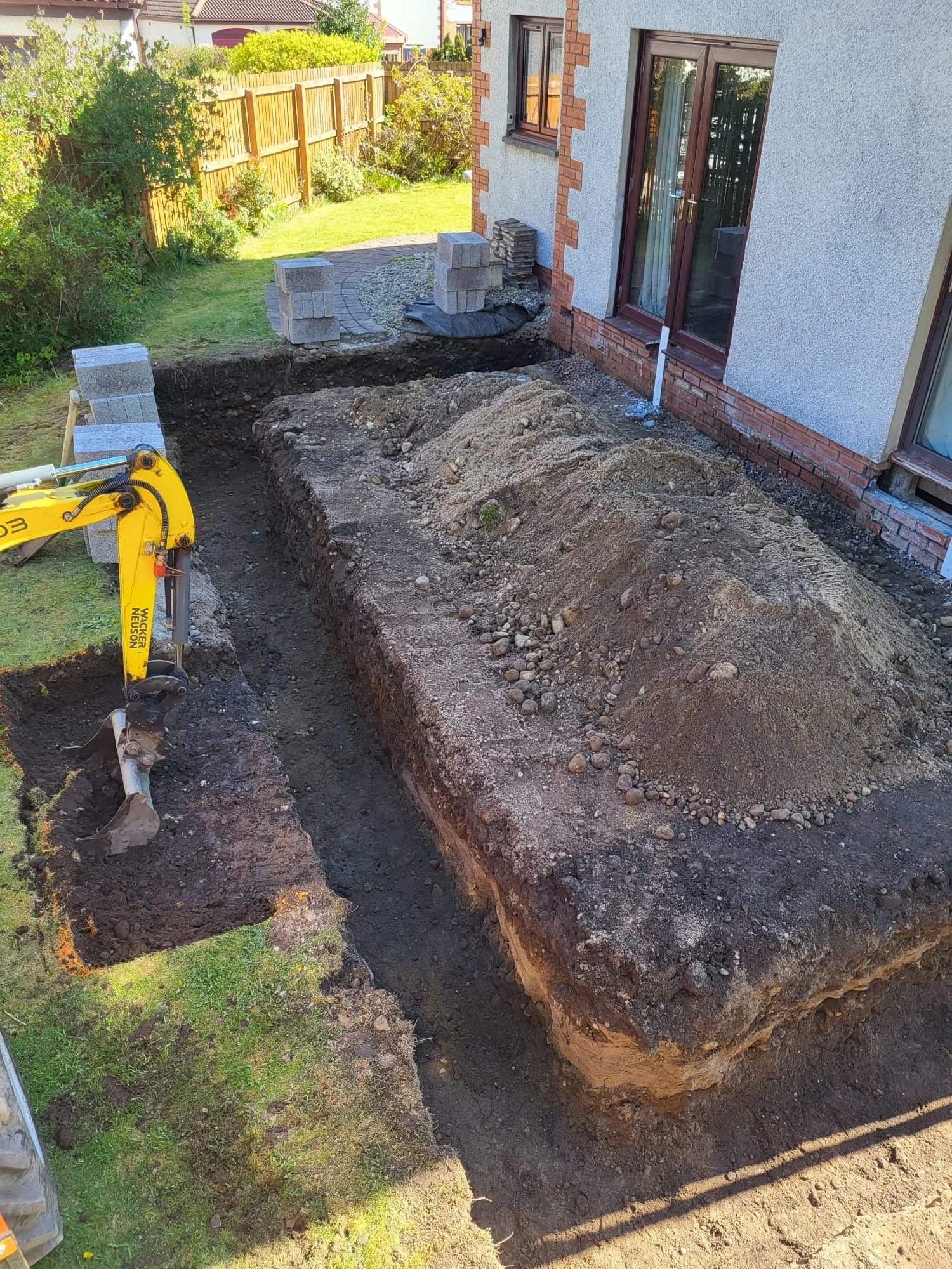 A backyard undergoing construction with a large trench dug into the ground. There are stone pavers, bricks, and construction materials near the house, which has a white exterior with brick accents. A small yellow excavator is visible in the lower lef