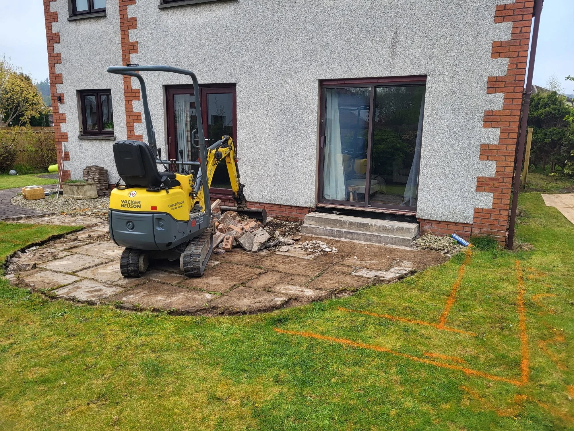 A house's exterior with ongoing construction work. A small yellow excavator is in front, working on the ground near the steps of a sliding glass door. The house has a textured white exterior with red brick accents and large windows. There are constru