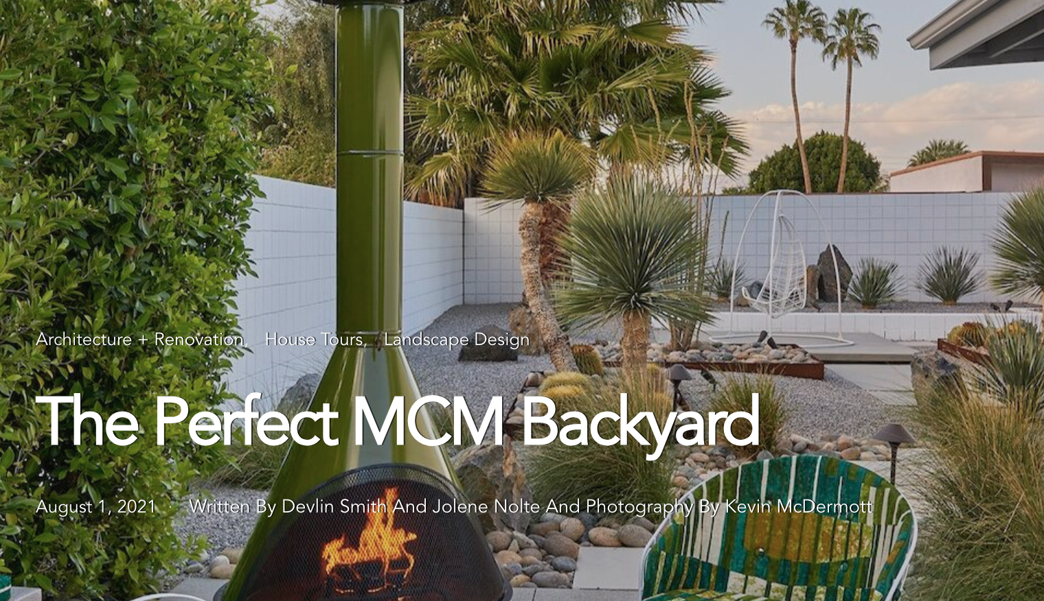 A backyard with desert plants, palm trees, gravel, and modern outdoor furniture, with a white fence and a fire pit in the foreground.
