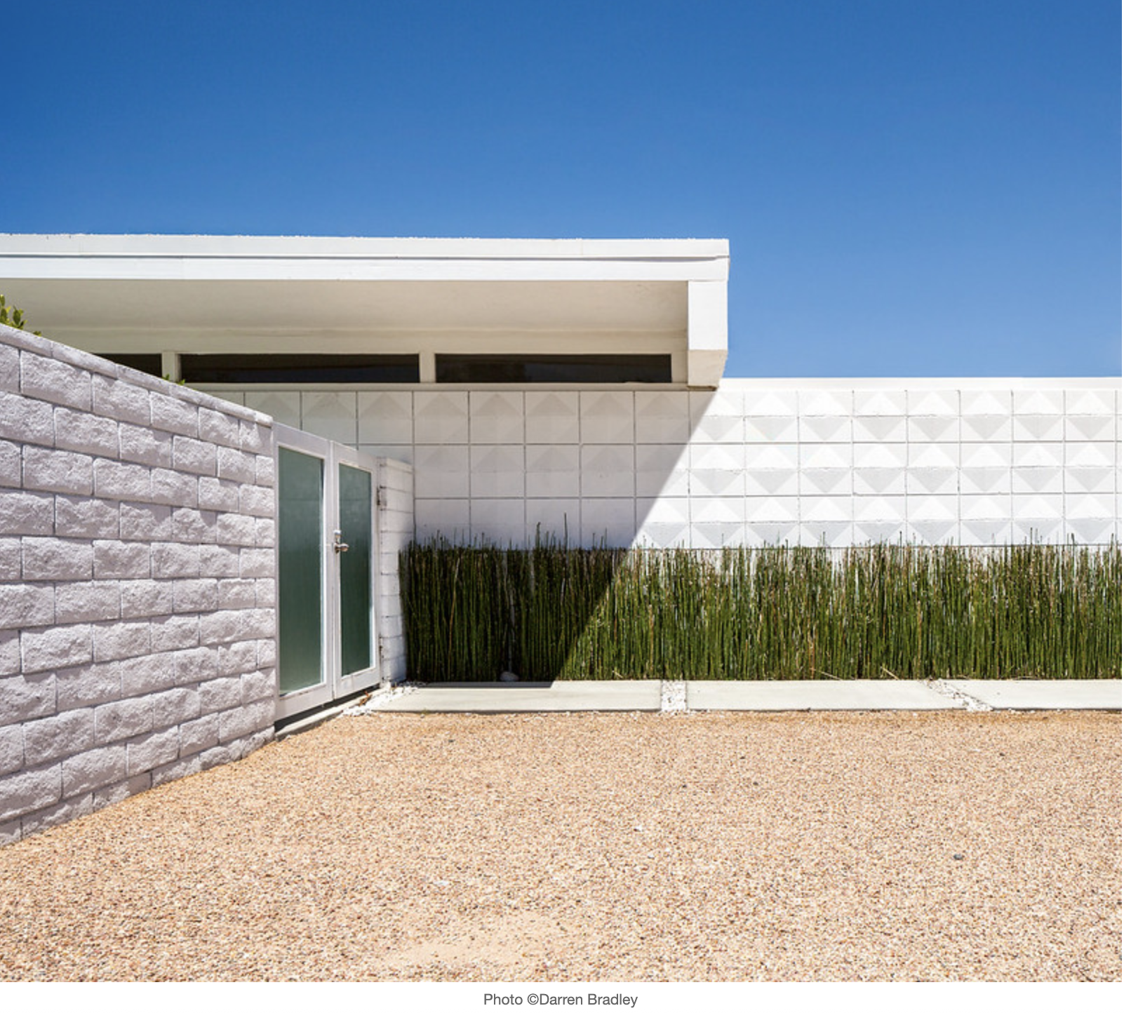 Modern house exterior with a white brick wall, a frosted glass door, and a garden with tall grass, set against a bright blue sky.