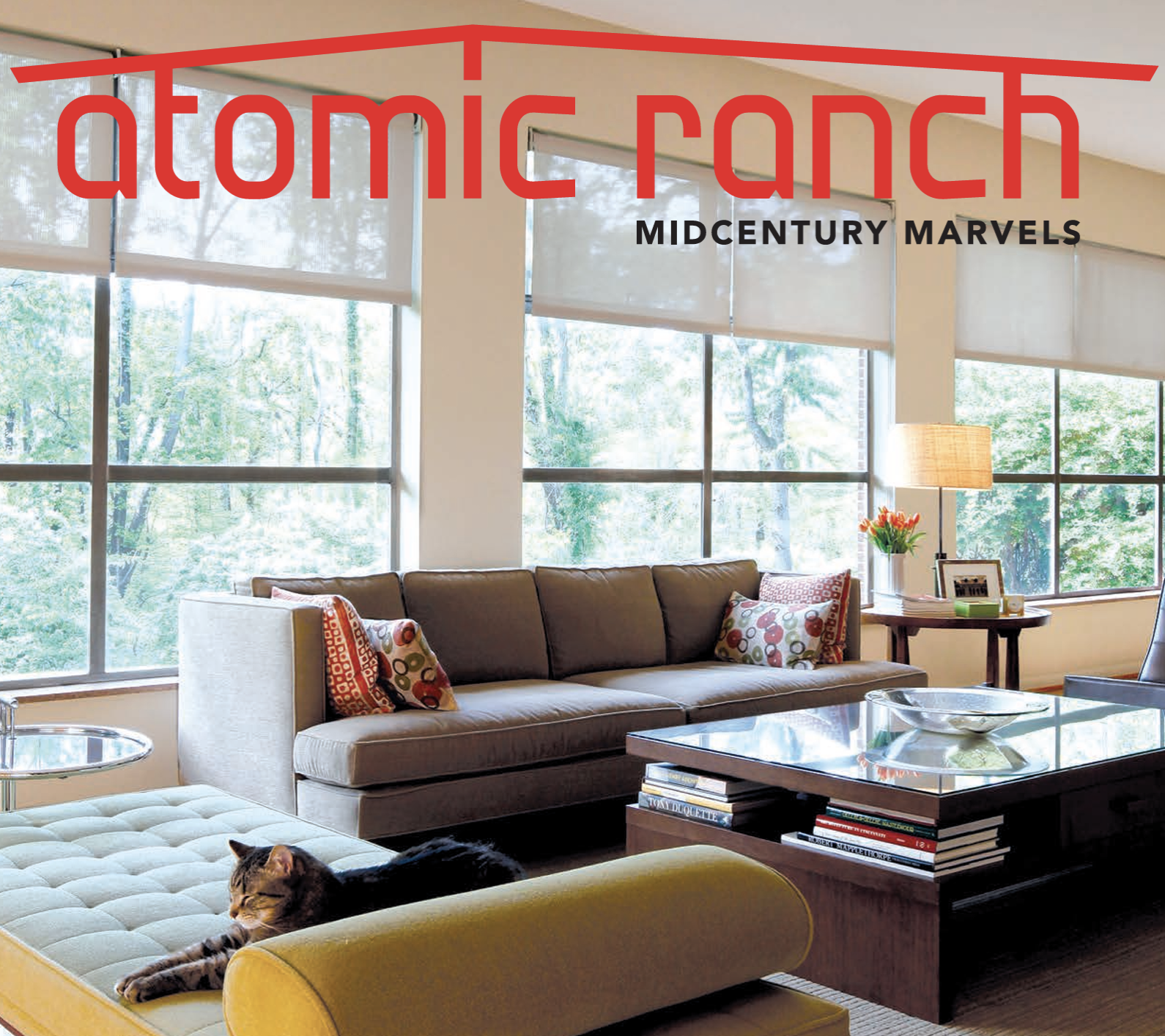 Living room with large windows, a brown sofa with colorful pillows, a yellow ottoman with a black cat, a wooden coffee table with books, a side table with a lamp, framed photo, and vase of orange flowers.