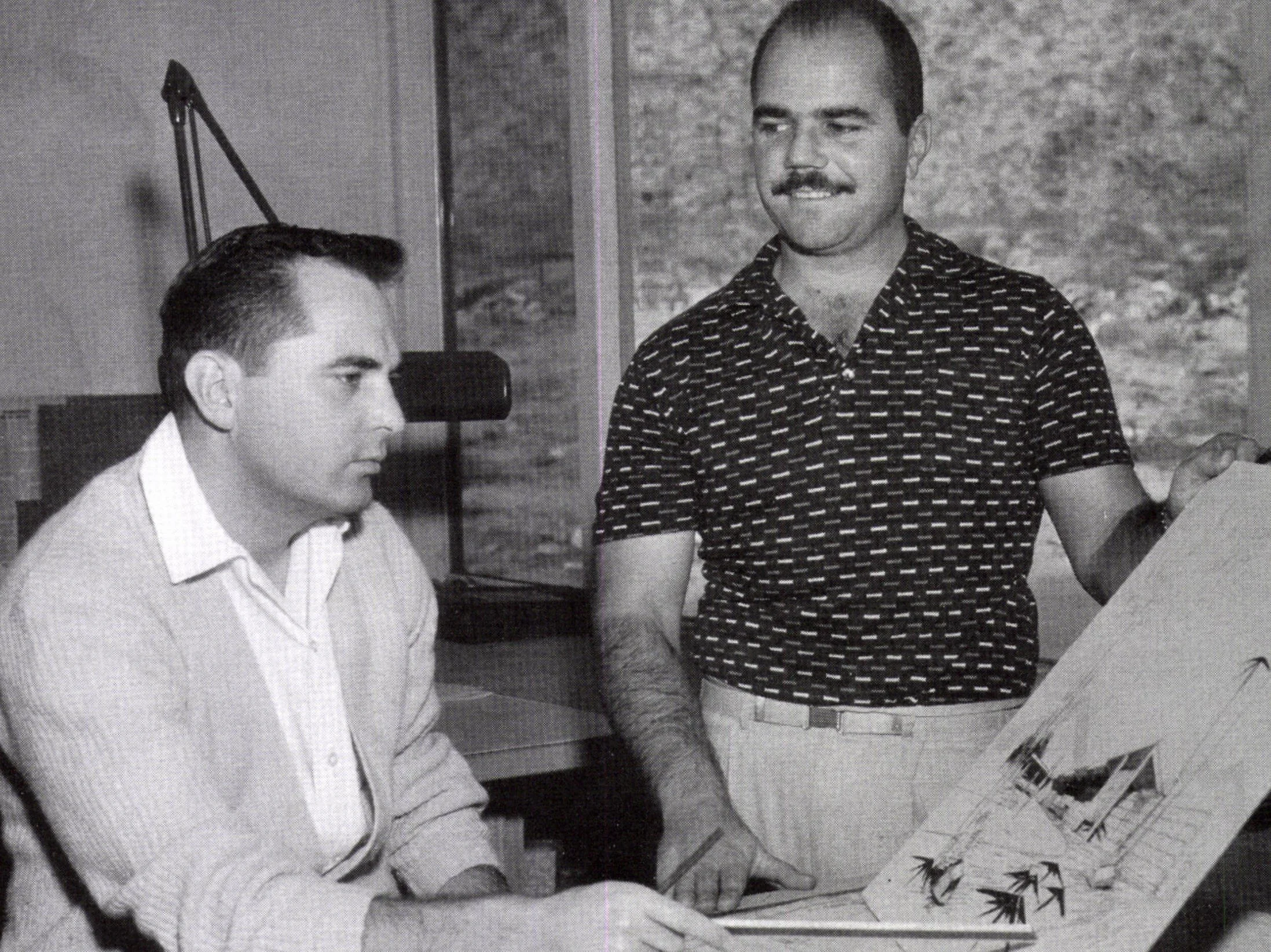 Two men in a discussion, one looking intently at documents on a table, the other standing and gesturing towards the papers, with a landscape painting in the background.