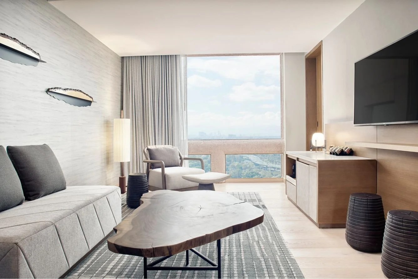 Modern hotel living room with large window overlooking city view, featuring a gray sofa with dark pillows, a marble-top coffee table, a white armchair, a wooden cabinet with a lamp, two black stools, and wall decor.