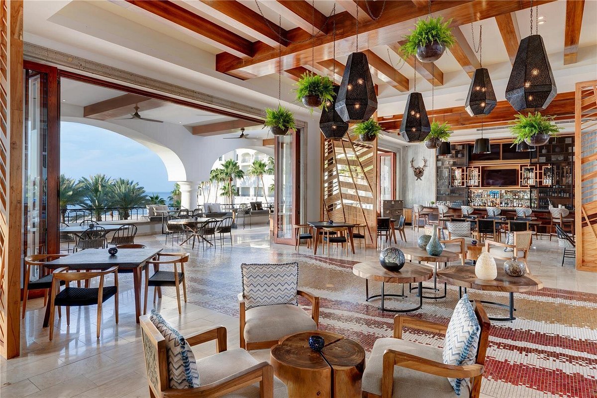 Elegant hotel lobby with wooden beams, hanging greenery, and modern pendant lights, featuring seating areas and an open view of a balcony with palm trees and the ocean.