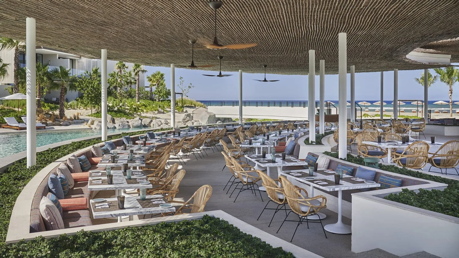 Open-air seaside restaurant with white marble tables, wicker chairs, ceiling fans, and ocean view in the background.