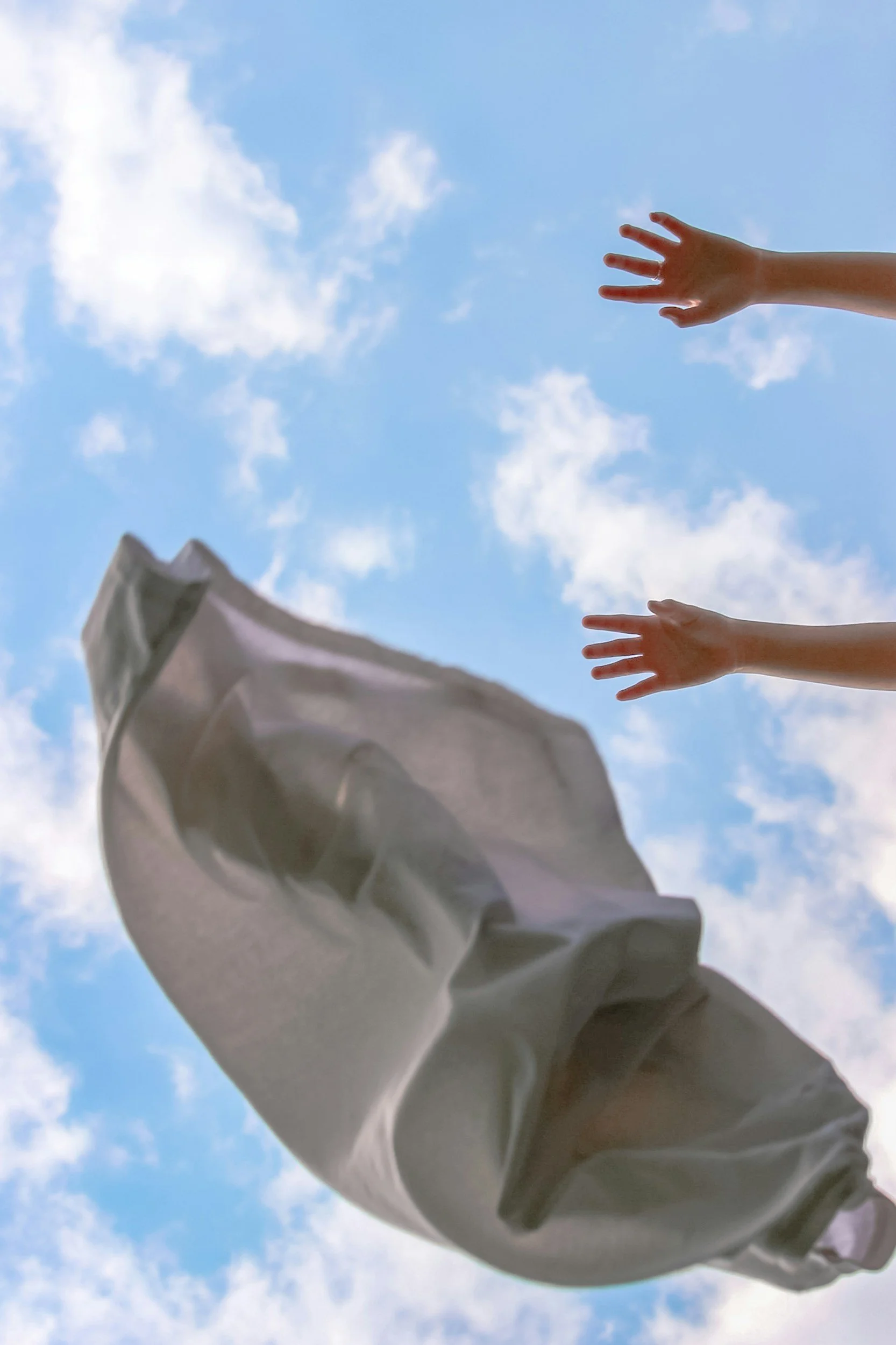 Two hands reaching out towards a bed sheet floating against a blue sky with clouds.
