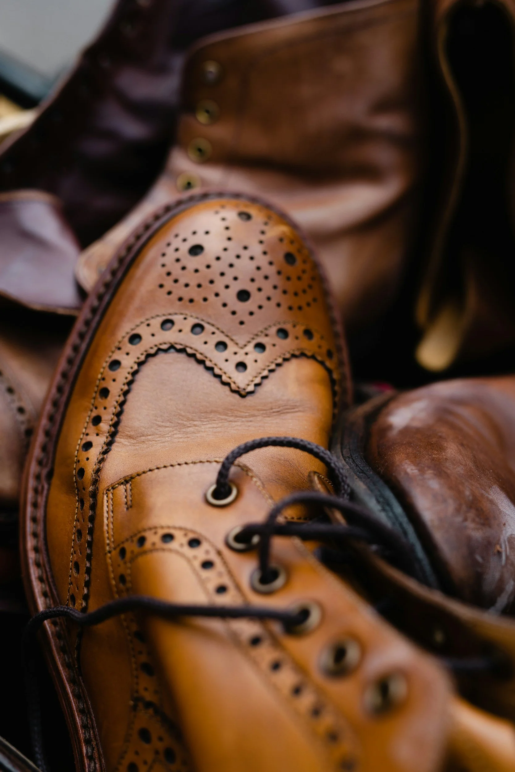 Close-up of a pair of tan leather brogue shoes with decorative perforations and dark laces, placed on top of darker leather shoes.