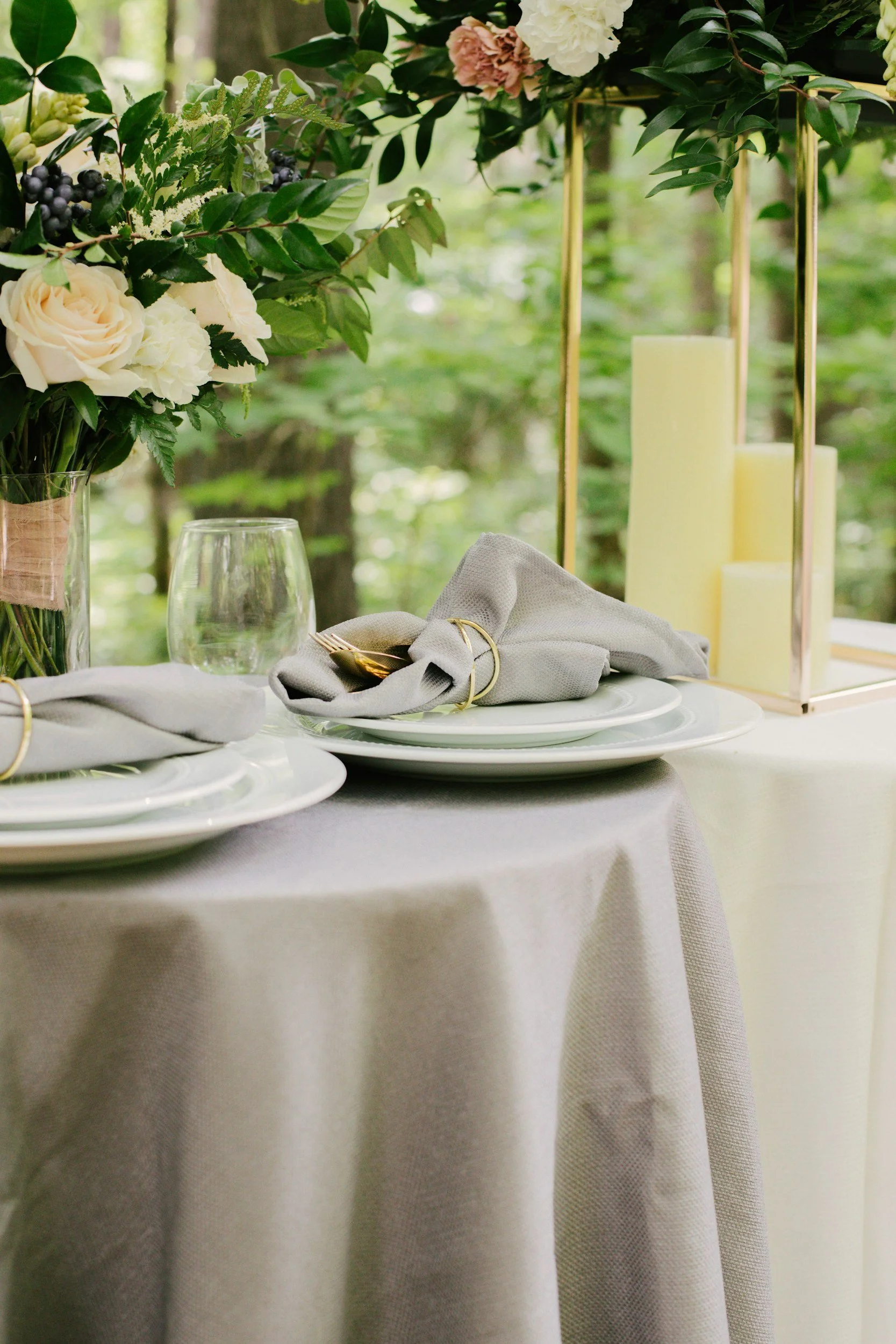 Elegant outdoor table setting with white plates, gray napkins with gold rings, clear glassware, a floral centerpiece with white and blush roses, greenery, dark berries, and three tall ivory candles inside a brass candle holder, against a backdrop of trees.