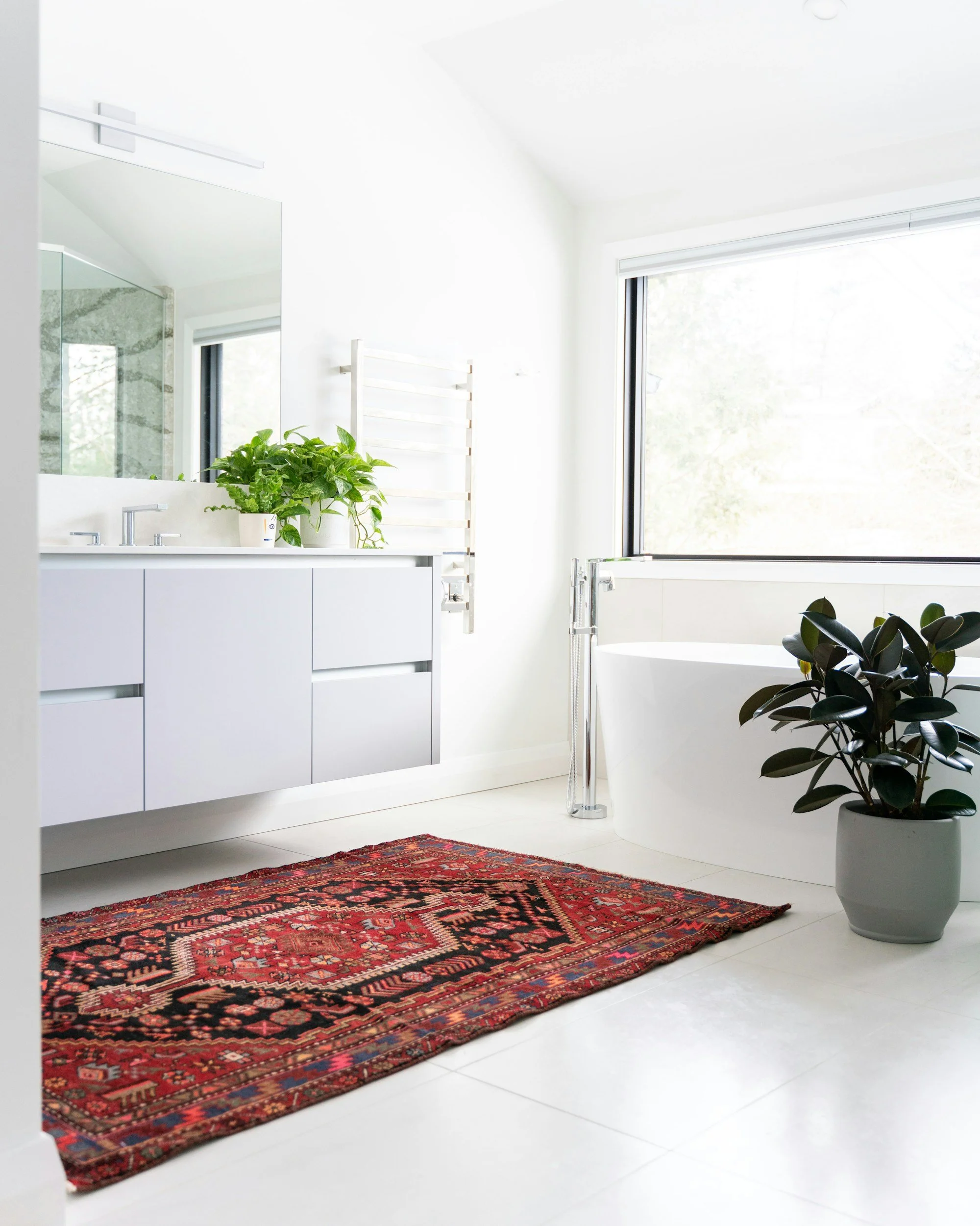 Bright bathroom with white vanity, large mirror, potted green plants, freestanding bathtub near window, red patterned rug on white tiled floor.