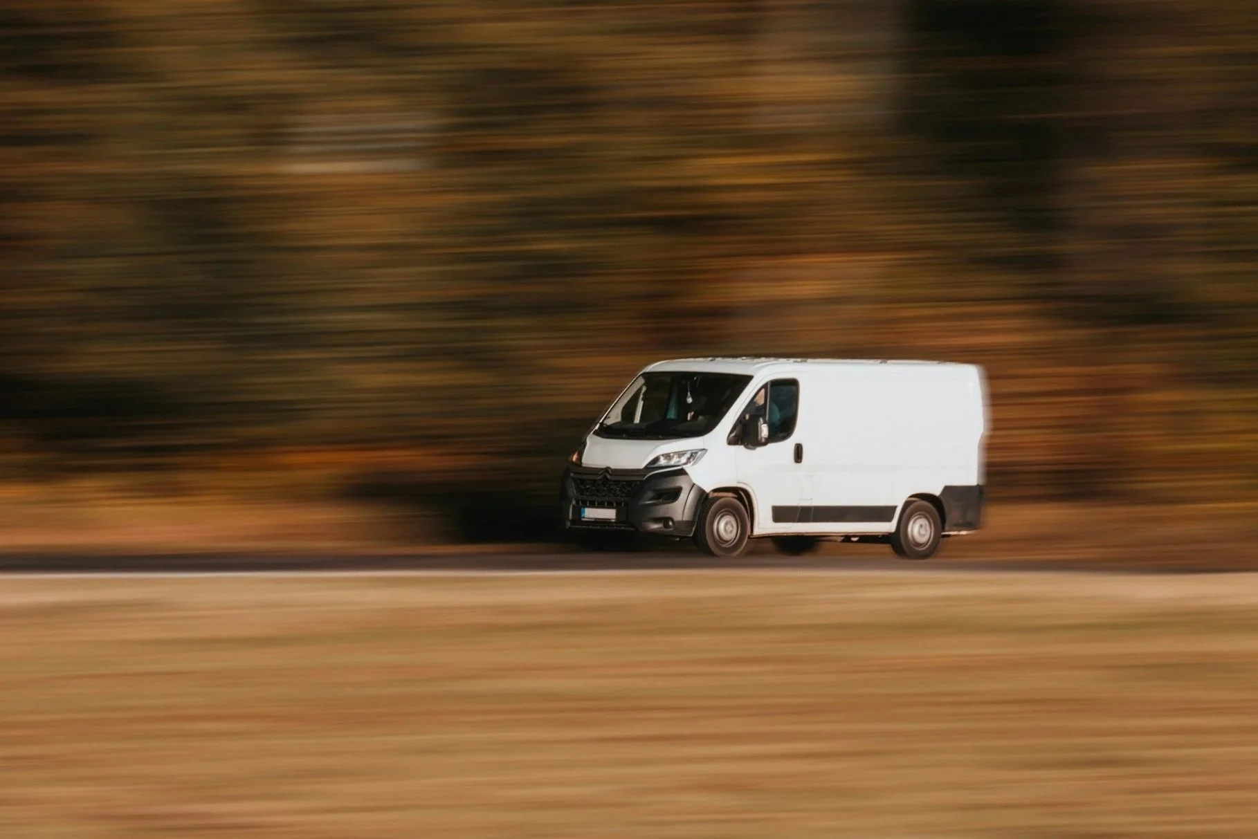 A white cargo van driving quickly along a road, with a blurred background of trees and landscape, suggesting motion.