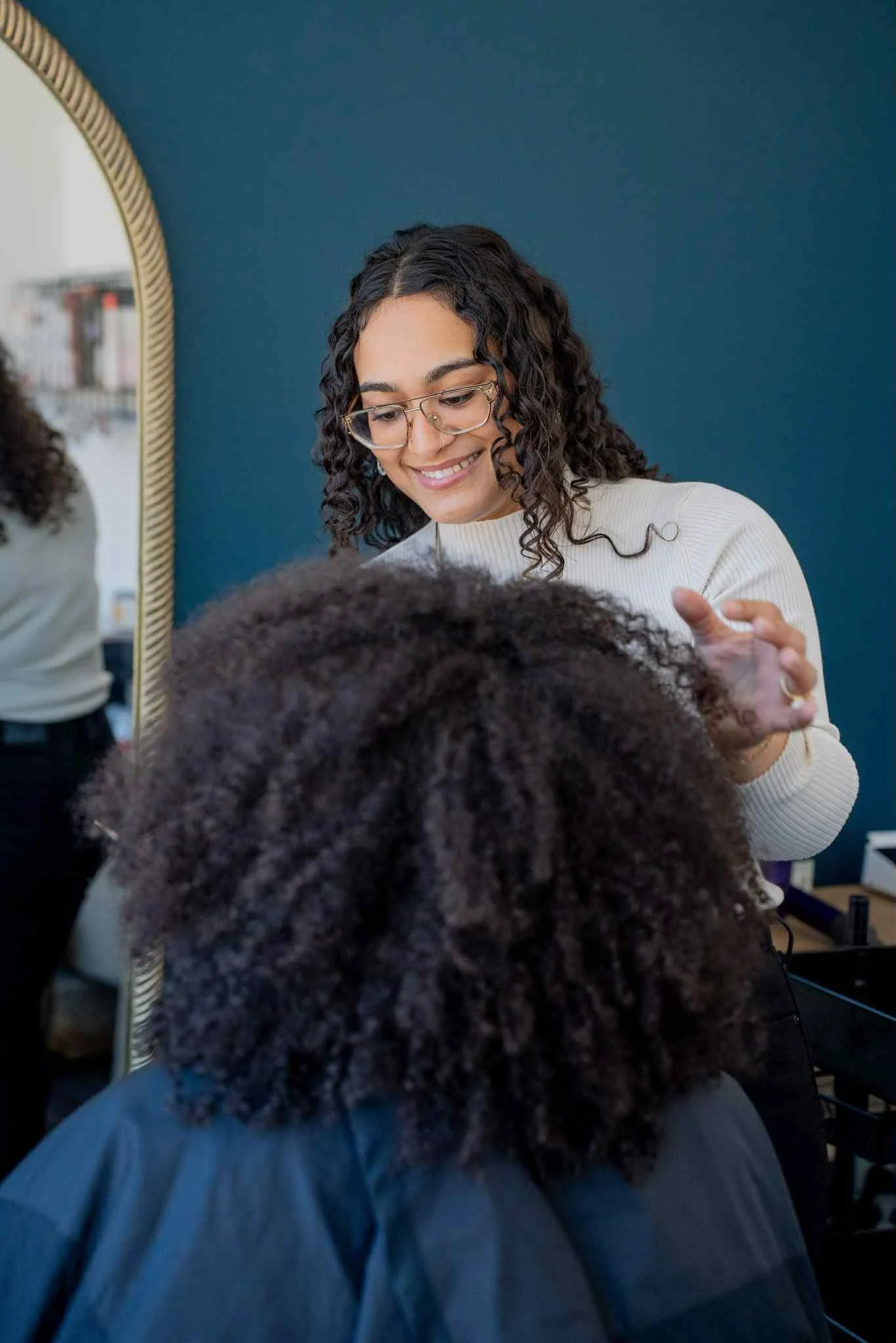 A hairstylist with glasses and curly hair smiling as she styles a client's natural curly hair.