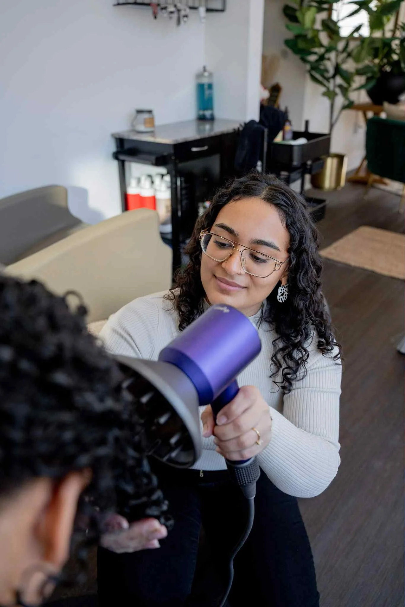 A woman with curly hair and glasses is using a purple hairdryer on another person's hair in a salon or similar setting.