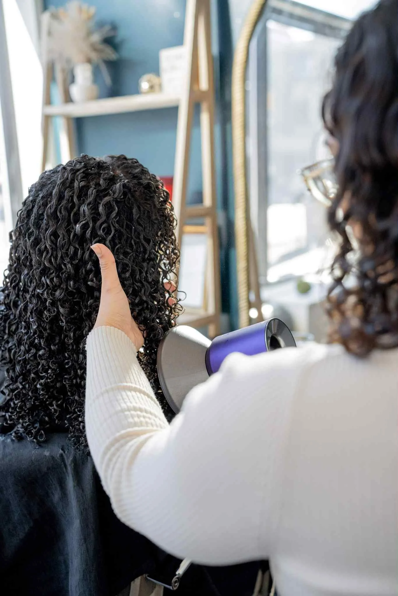 A hairstylist is using a purple blow dryer to dry a woman's curly hair in a salon. The woman with curly hair is seated, and the stylist is holding the dryer with one hand while supporting the woman's head with the other. The salon has a large window, a shelf with decor, and a blue wall in the background.