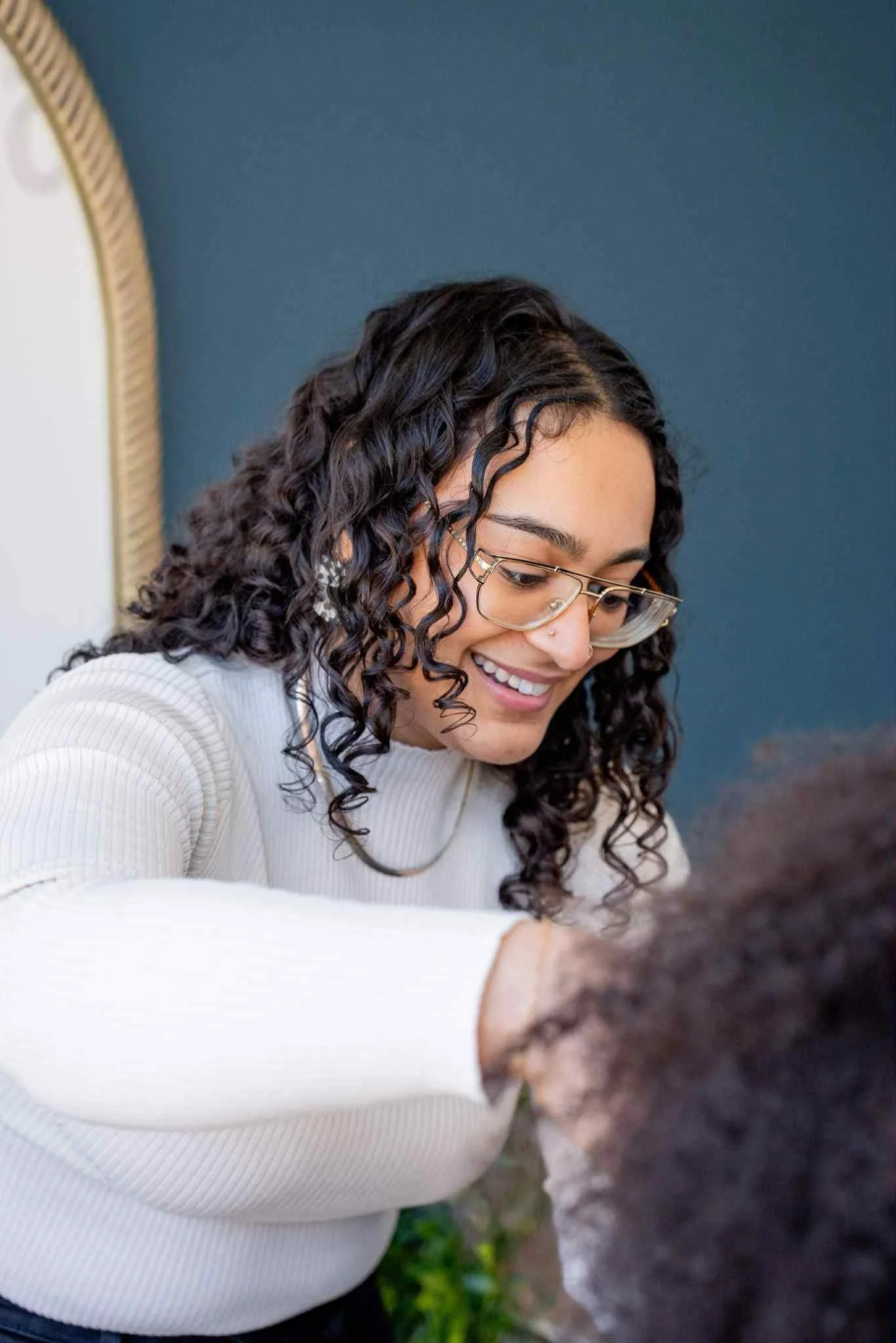 A woman with curly dark hair, glasses, and a nose piercing smiling while looking down.