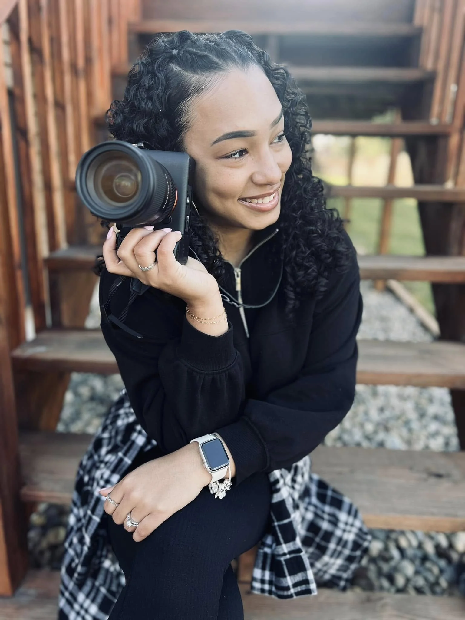 A woman with curly black hair sitting on outdoor wooden stairs, smiling while holding a camera to her face.