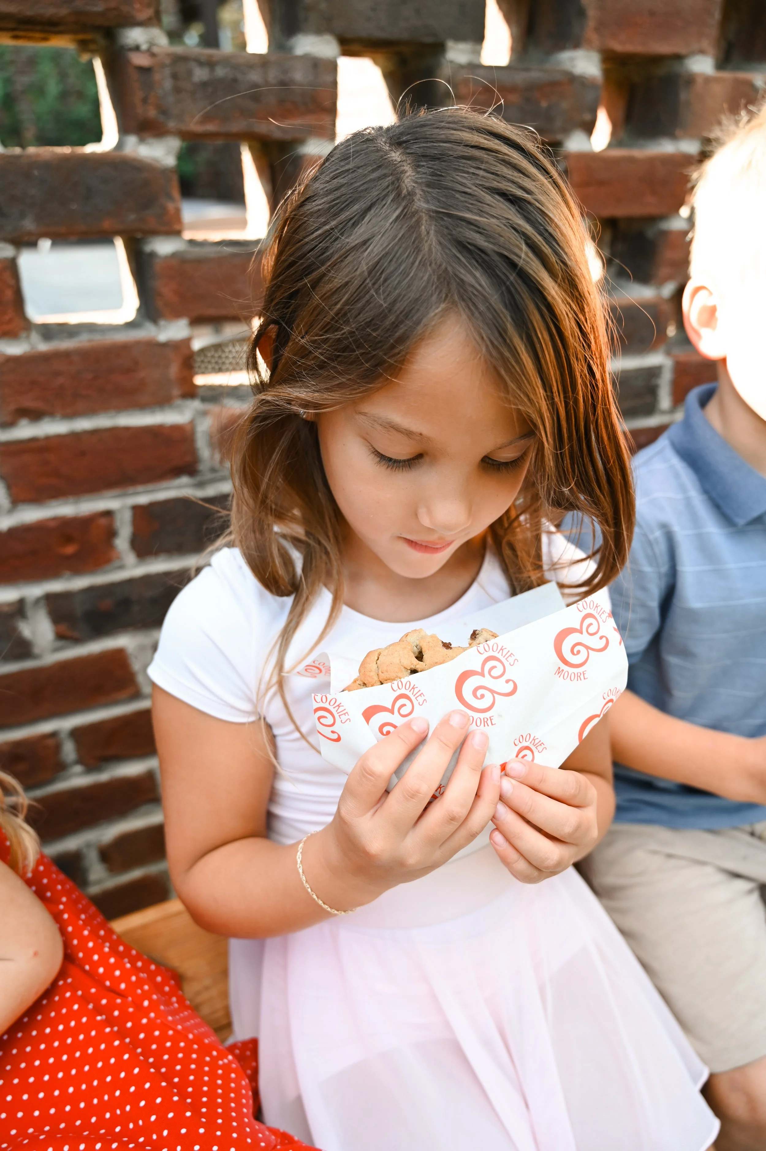 Girl in white dress holding a cookie in a paper wrapper with red spiral logo, sitting next to children, against a brick wall background.