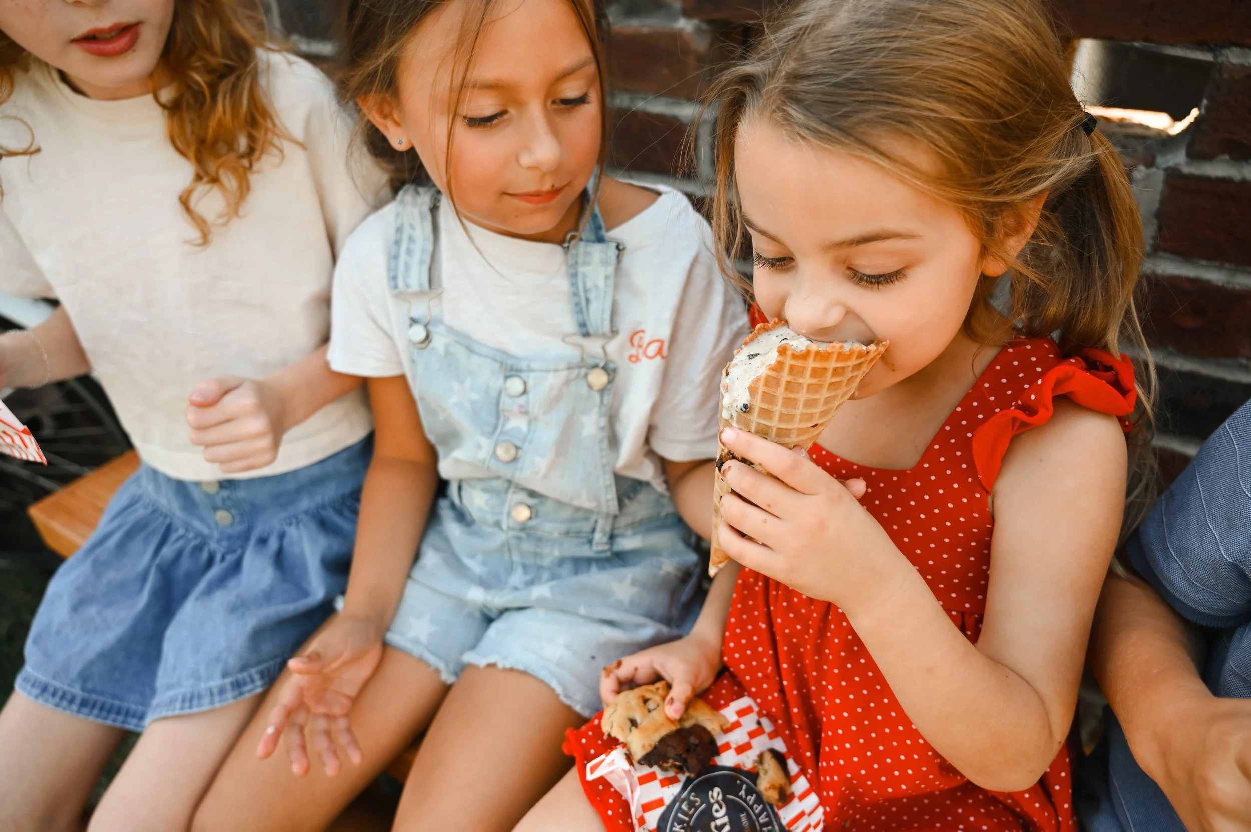 Girl in red polka dot dress enjoying ice cream cone with other children sitting beside her.