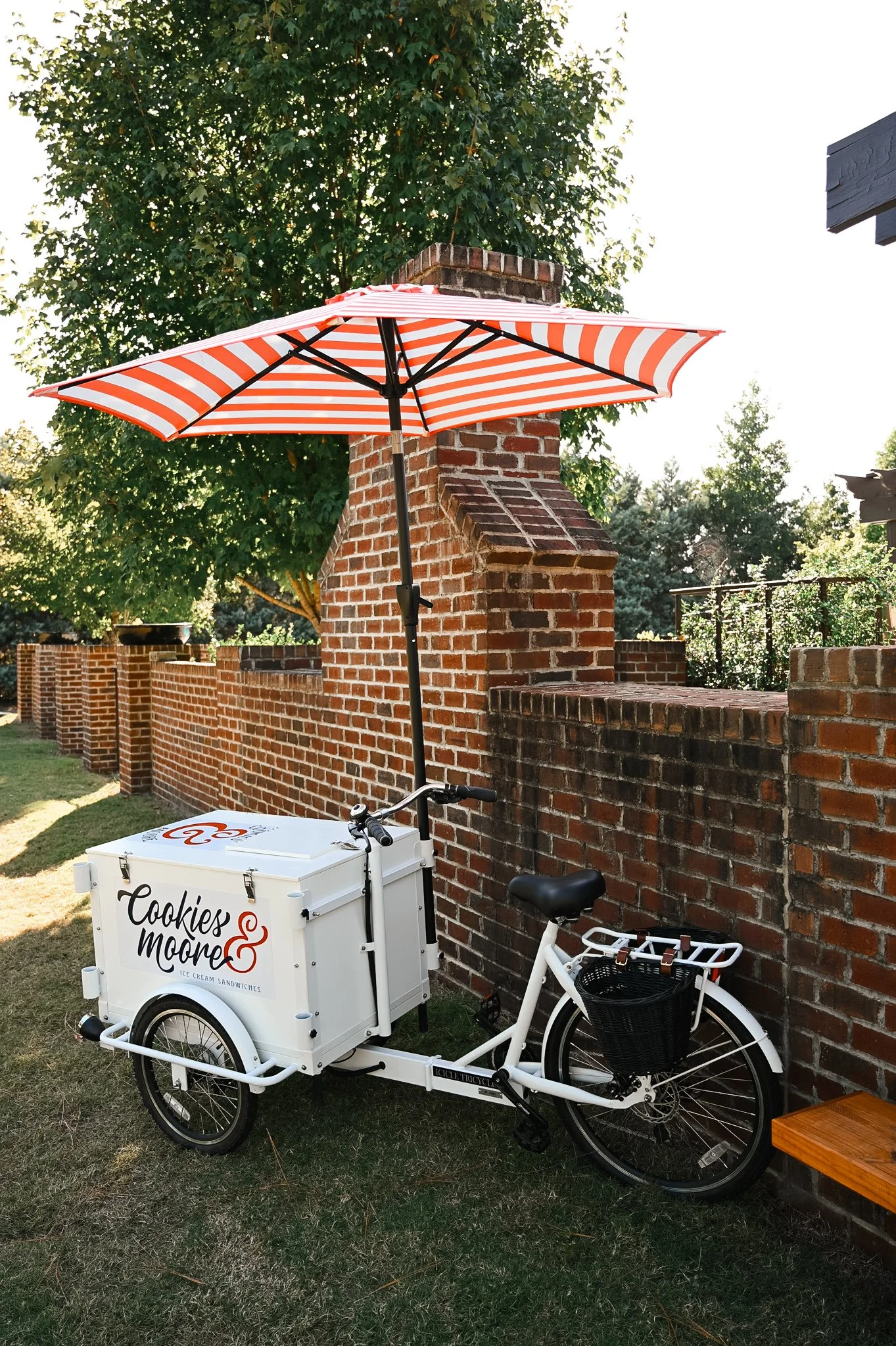 The Cookies & Moore Ice Cream Bicycle, branded with "Cookies & More Ice Cream Sandwiches", parked next to a brick wall with a red and white striped umbrella attached to it for shade.