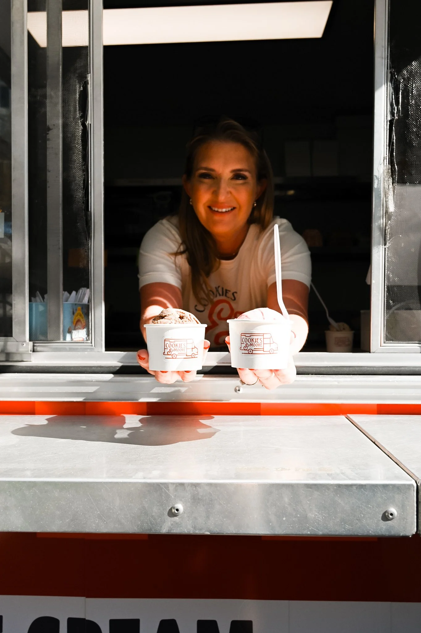 A woman in a white t-shirt with a logo, smiling and holding two cups of ice cream through a food truck window.