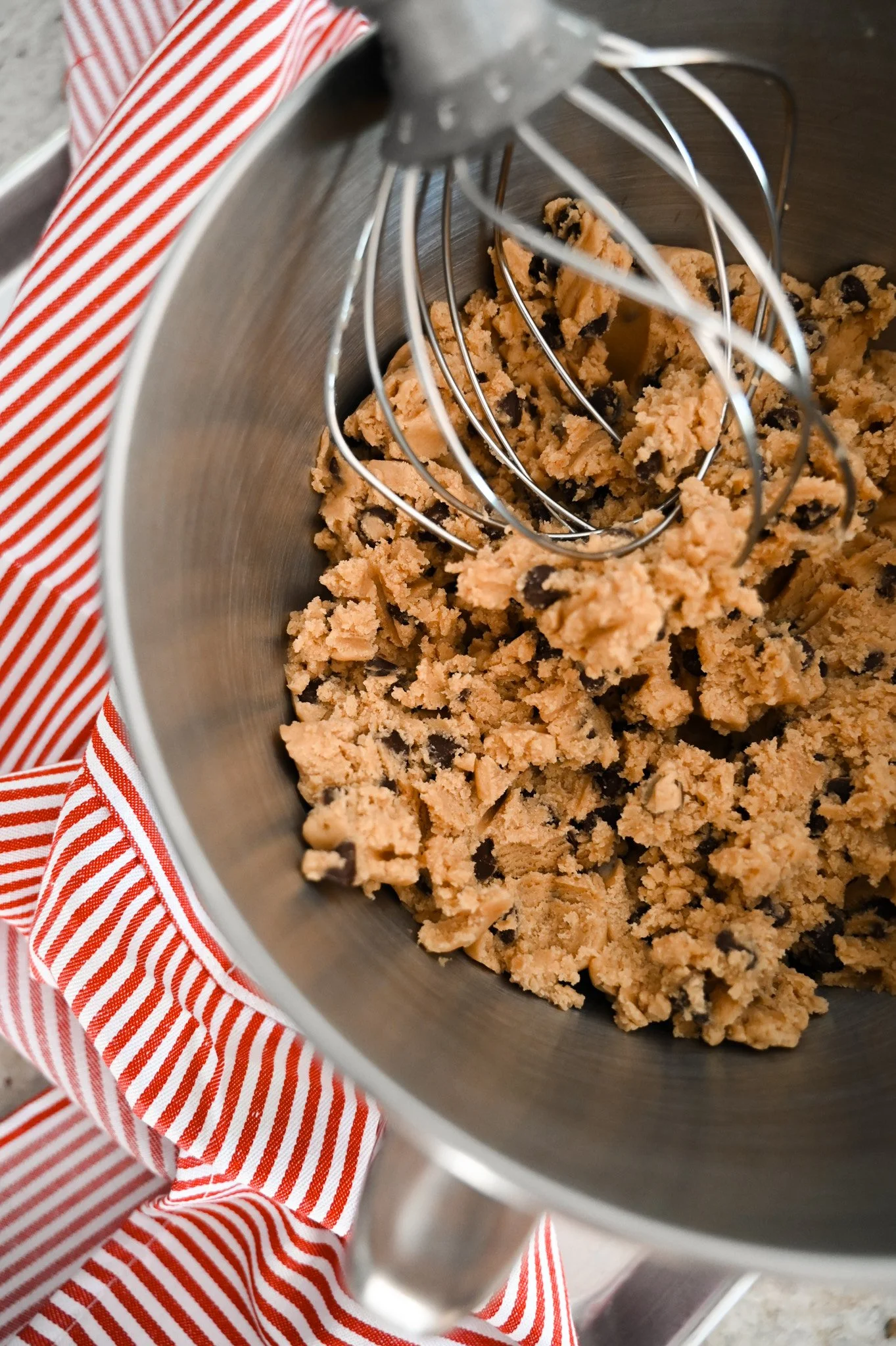 Chocolate chip cookie dough in a mixing bowl with a whisk attachment, red and white striped cloth underneath.