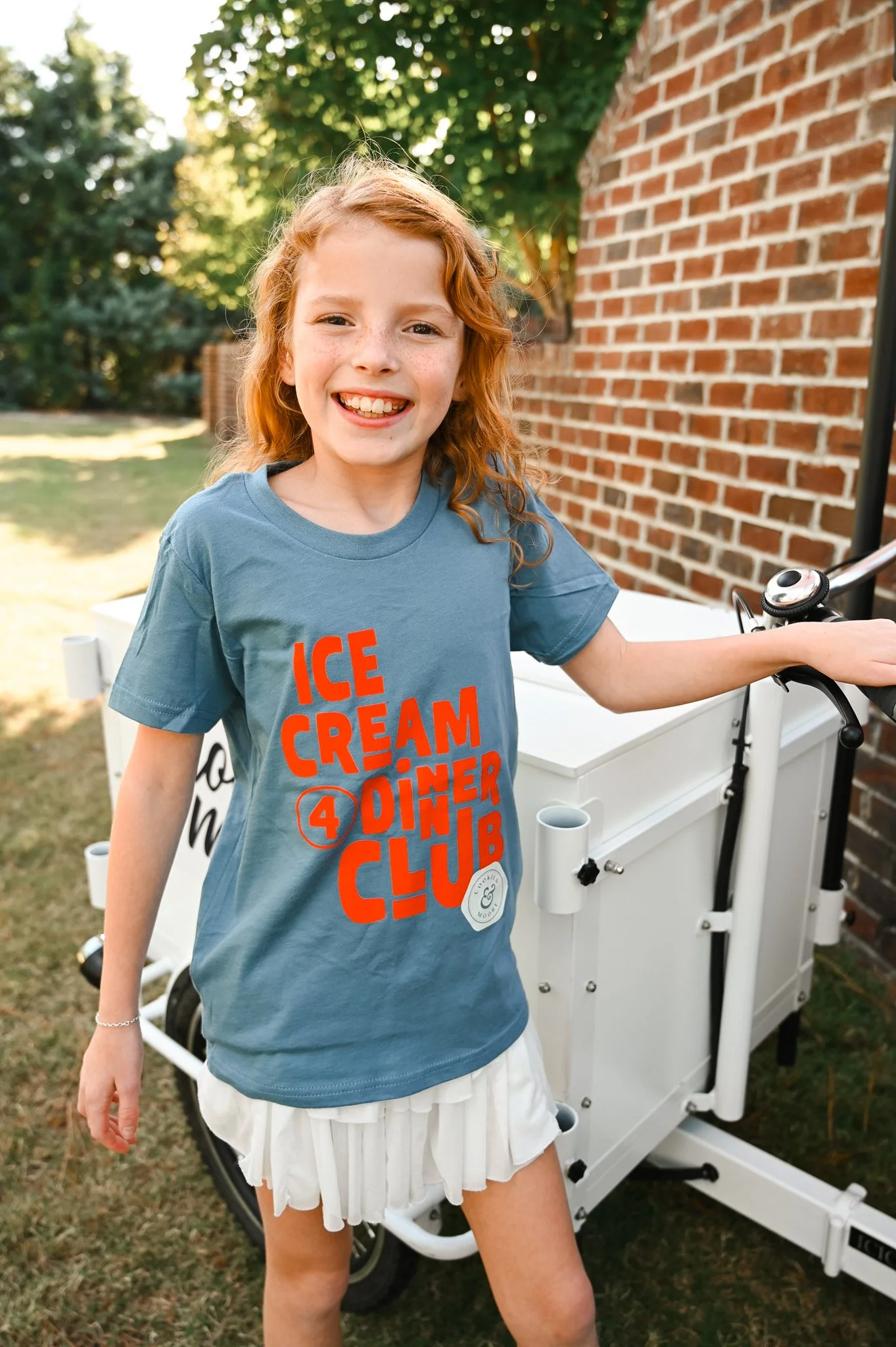 A young girl with red hair and freckles smiling outdoors, wearing a blue t-shirt with red text that says 'Ice Cream 4 Dinner Club' and a white skirt, standing next to a white ice cream cart.