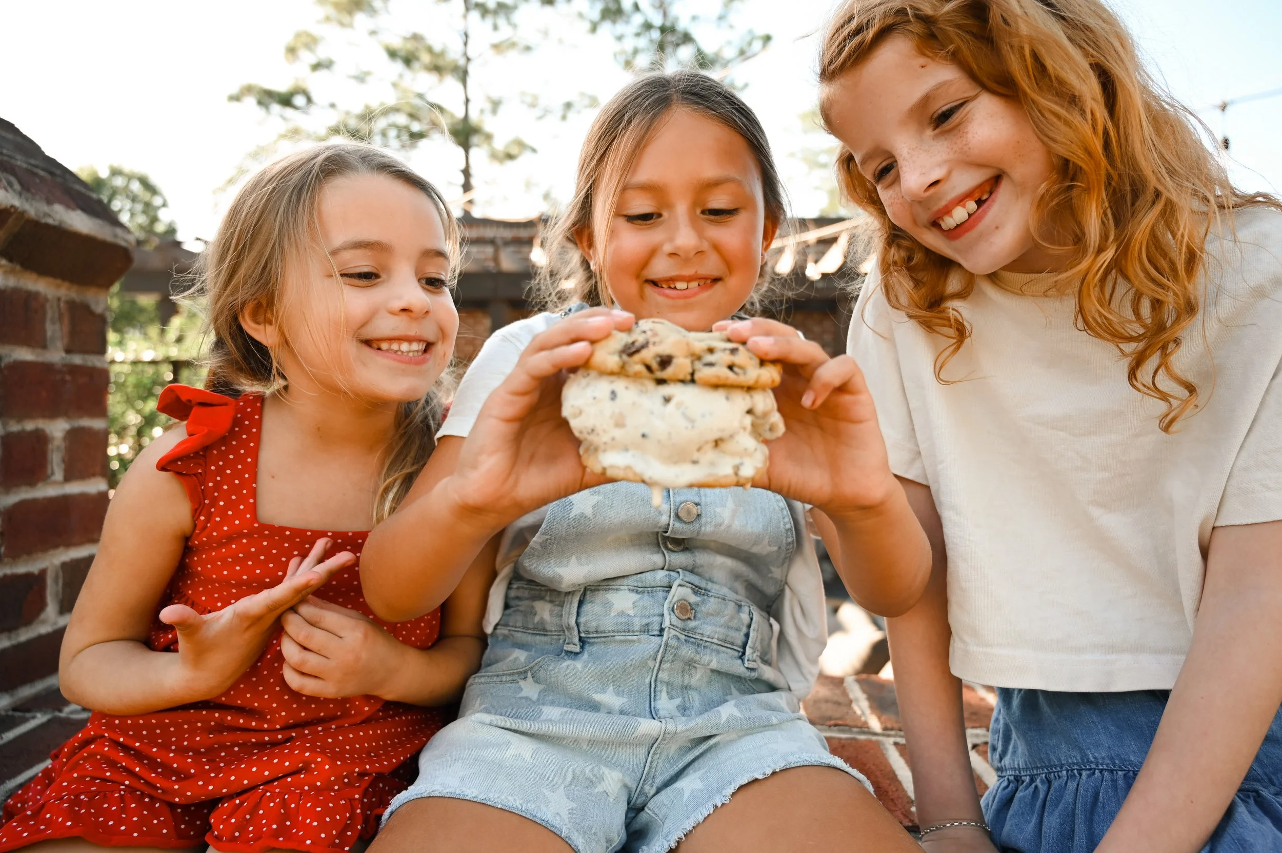 Three young girls sitting outdoors, smiling, as one girl holds a large cookie and ice cream sandwich from cookies & Moore, with a brick wall and trees in the background.