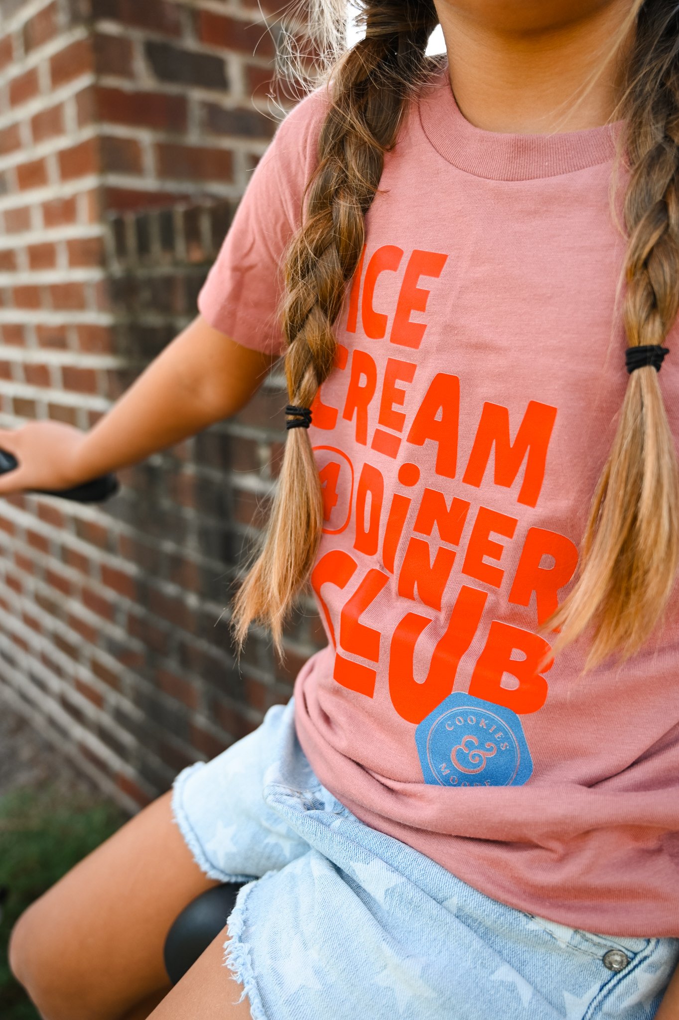 Close-up of a girl with braided hair wearing a pink t-shirt with red text that reads 'Ice Cream 4 Dinner Club and light blue denim shorts, sitting outdoors near a brick wall.