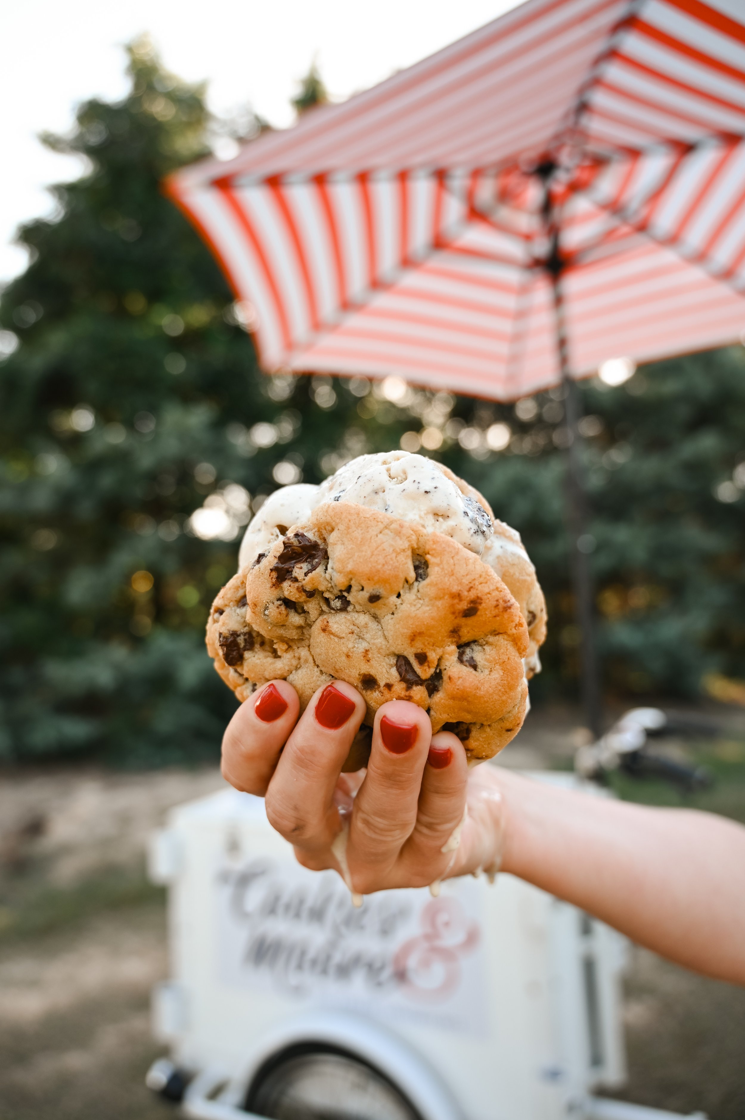 Person holding three large cookies with ice cream scoops in the background, red outdoor umbrella, and an ice cream cart in a park.