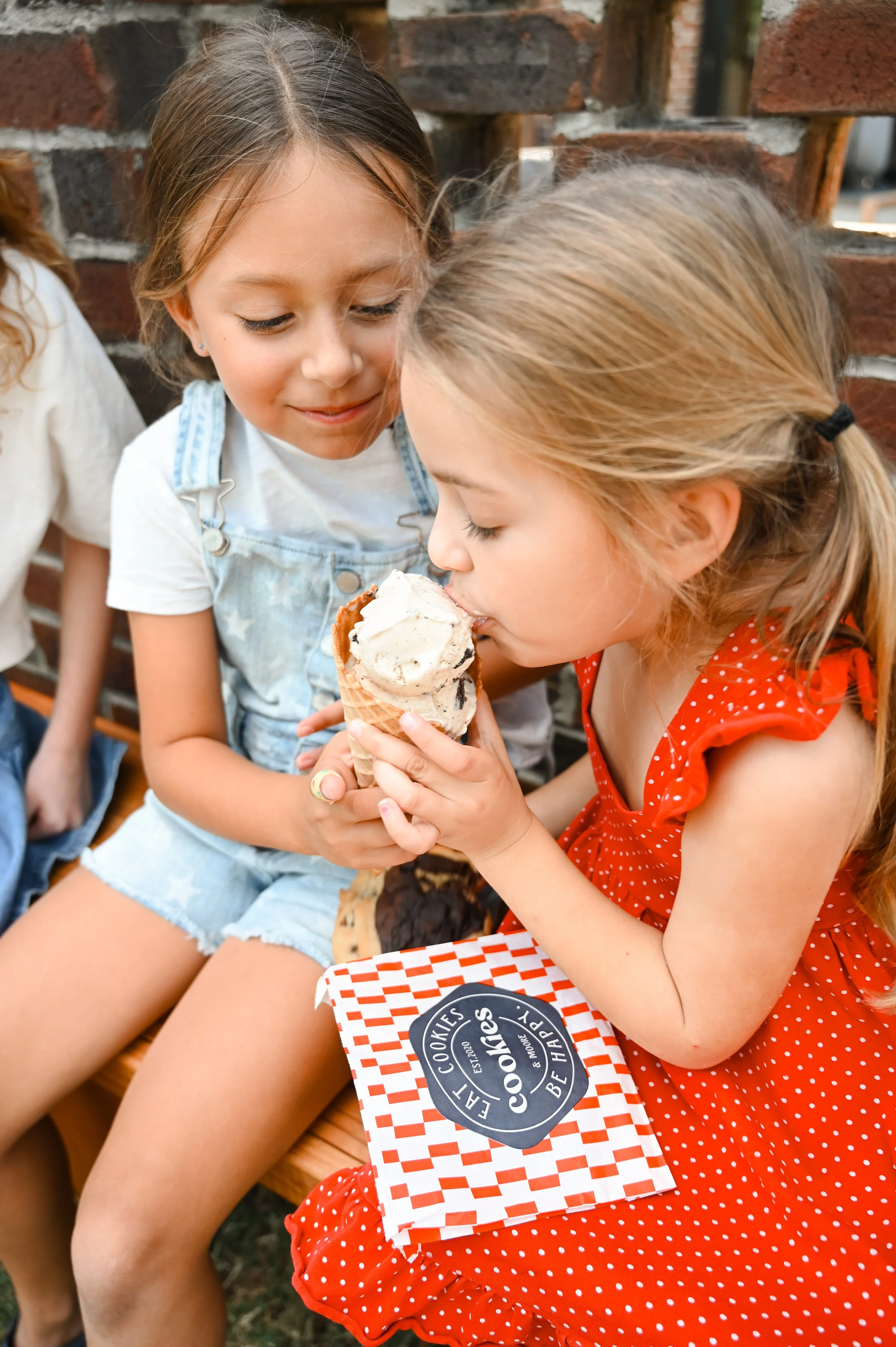 Two young girls sharing and tasting ice cream cone together outside, sitting near a brick wall. One girl is wearing a red polka dot dress, and the other girl is in light blue overalls and a white shirt.