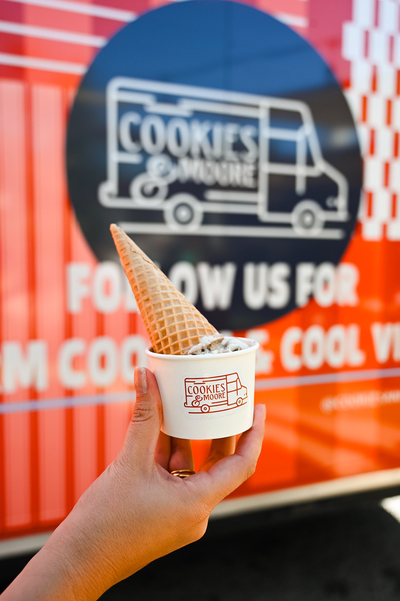 Person holding a cup of ice cream with a waffle cone piece on top, in front of a red and black sign for Cookies & Moore food truck.