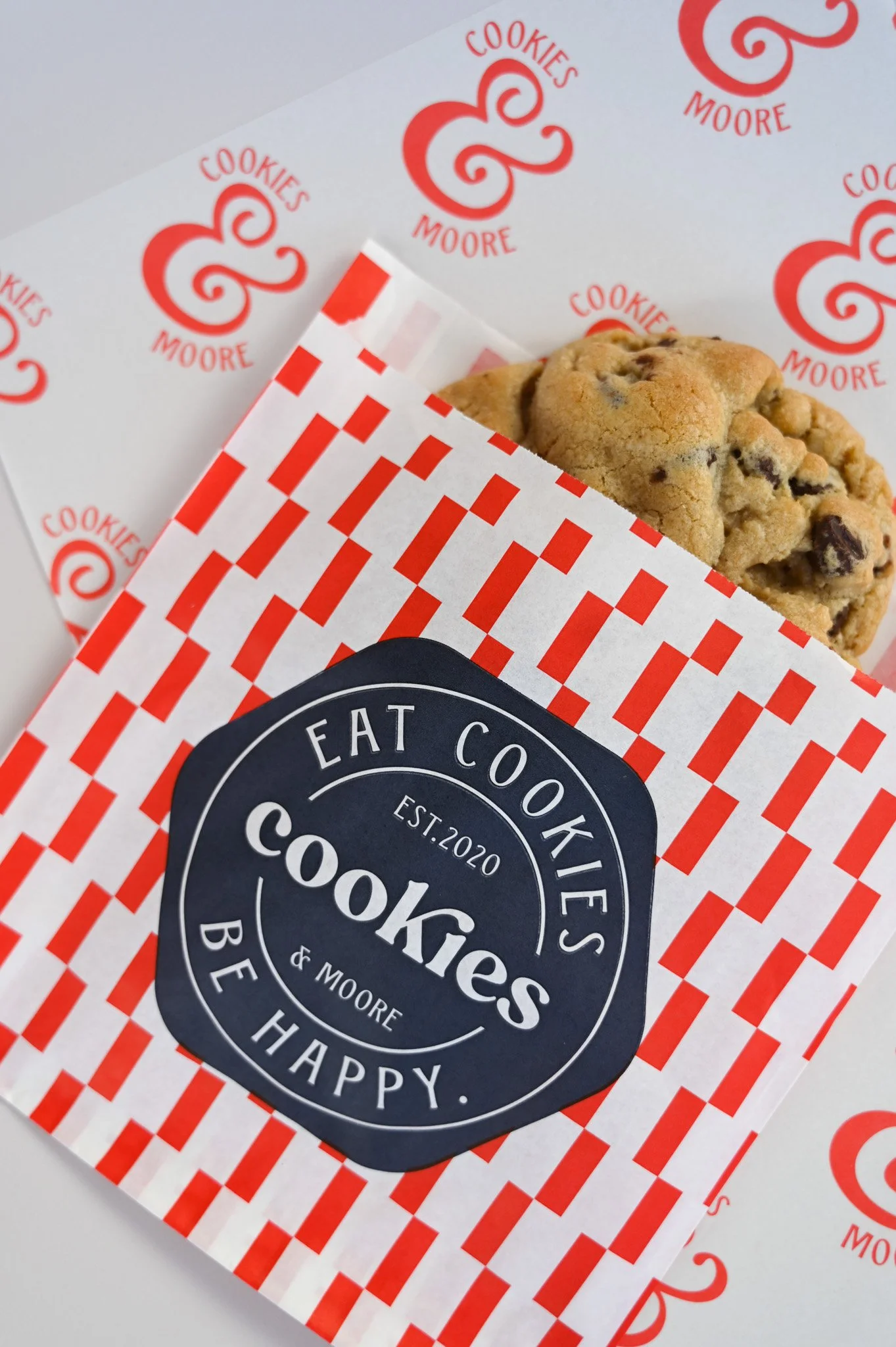 A chocolate chip cookie in a paper bag with a red and white checkered pattern, placed on top of a sheet of paper with 'Cookies & Moore' logo.