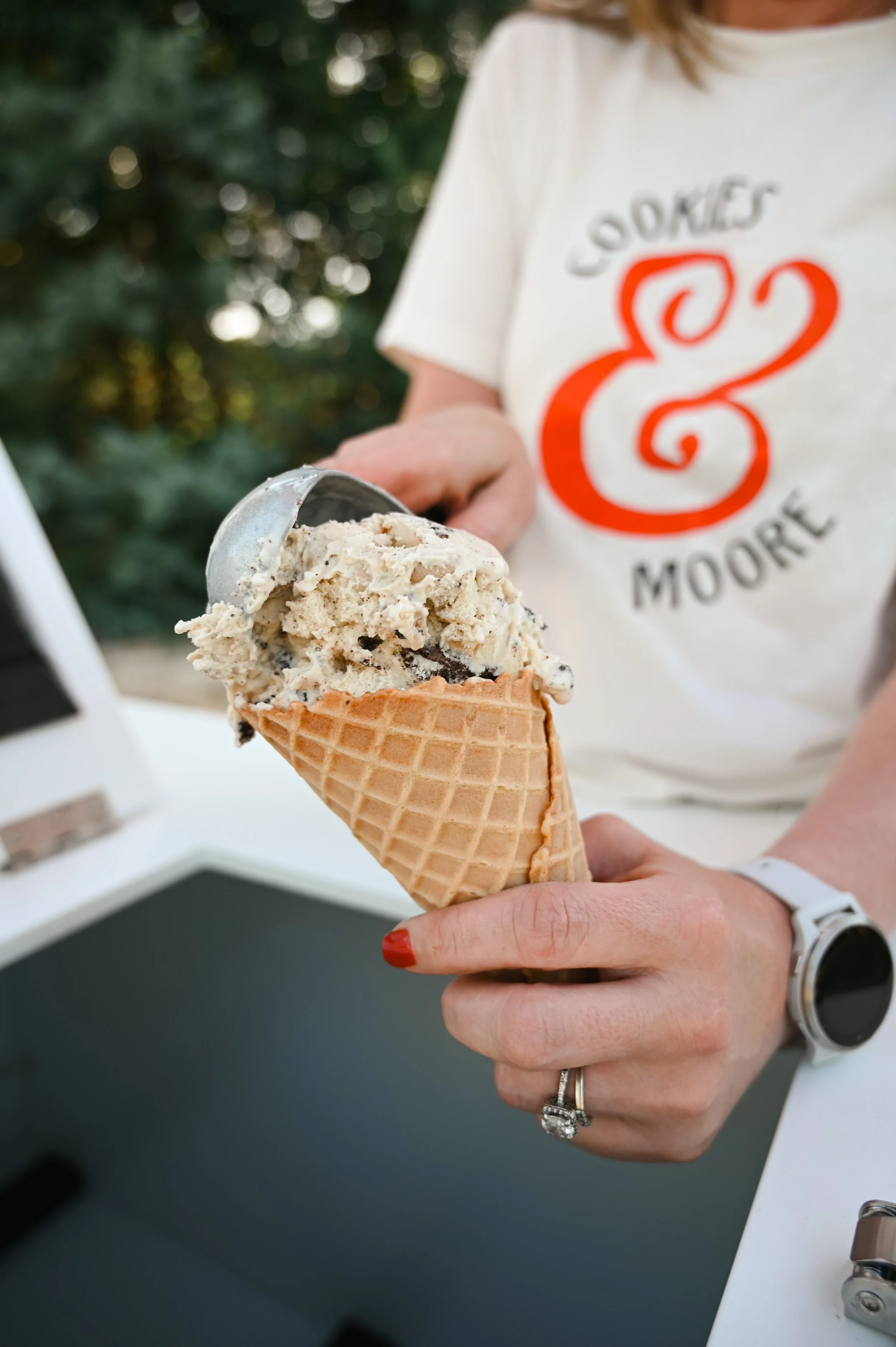 Person holding a waffle cone with scoop of cookies and cream ice cream, wearing a white t-shirt with red and black lettering, outdoor setting.