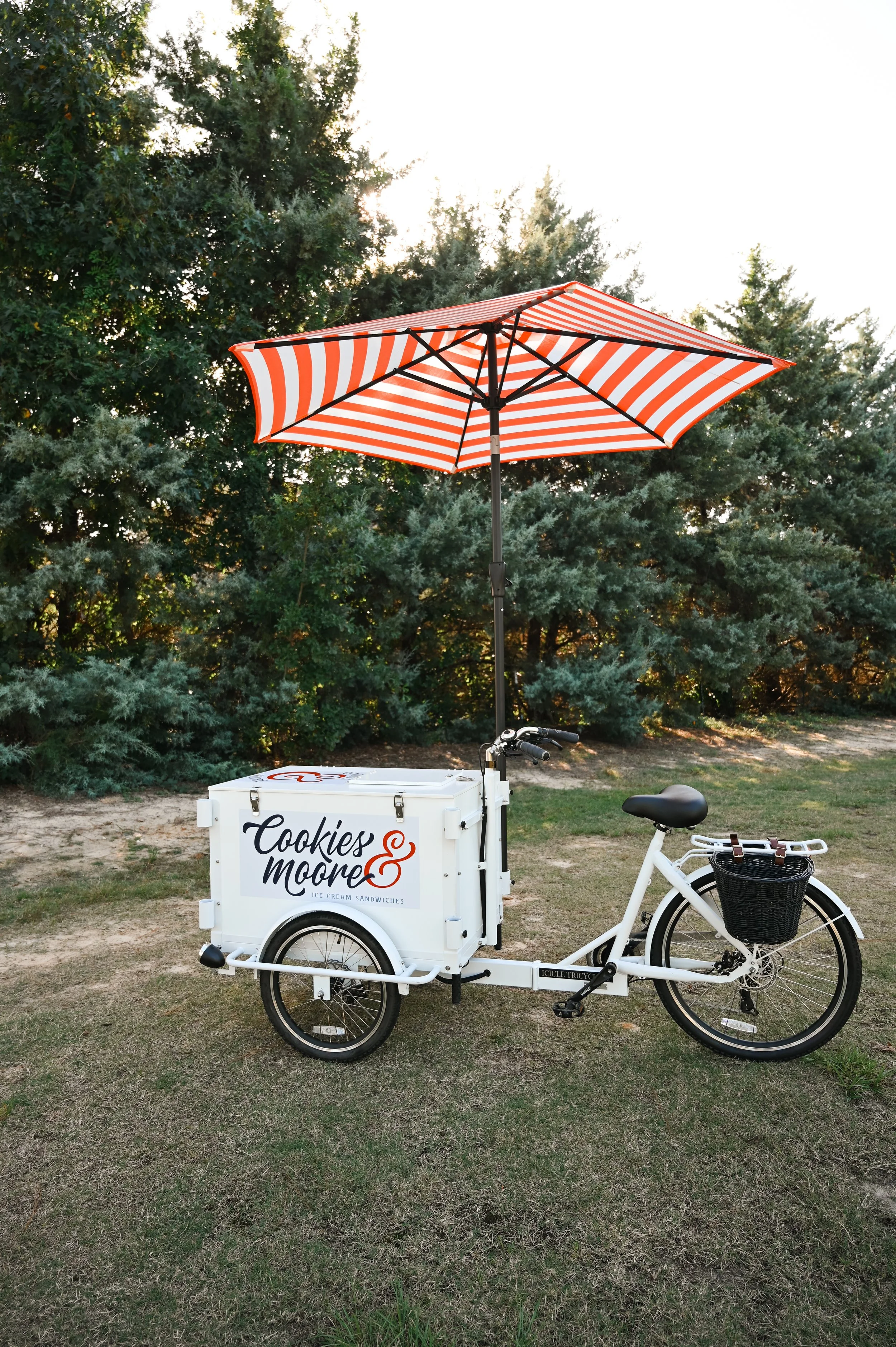 A white Ice bicycle selling Cookies & Moore ice cream sandwiches, equipped with a red and white striped umbrella attached above, outdoors on grass with trees in the background.