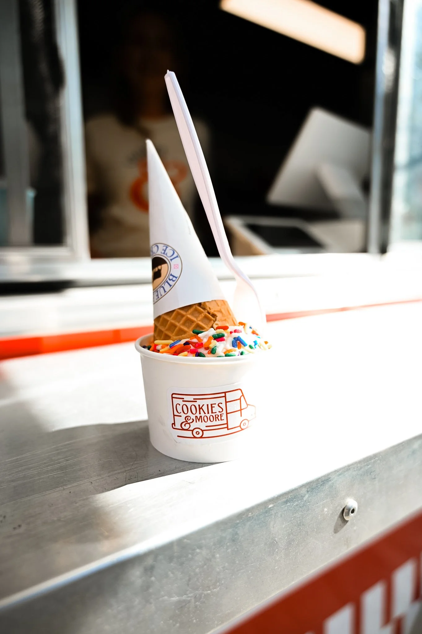 A cup of vanilla ice cream with rainbow sprinkles and a waffle cone inside, with a pink spoon, on a metal counter at the Cookies & Moore Food Truck in Moore County, North Carolina.