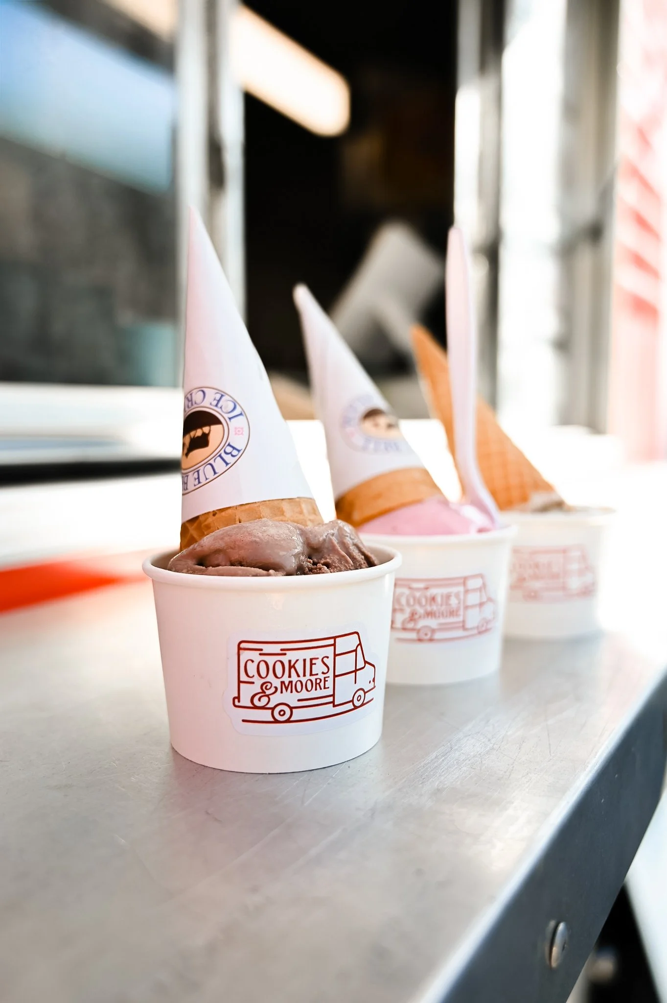 Three cups of ice cream with cone hats on a metal surface, with Cookies & Moore branding visible.