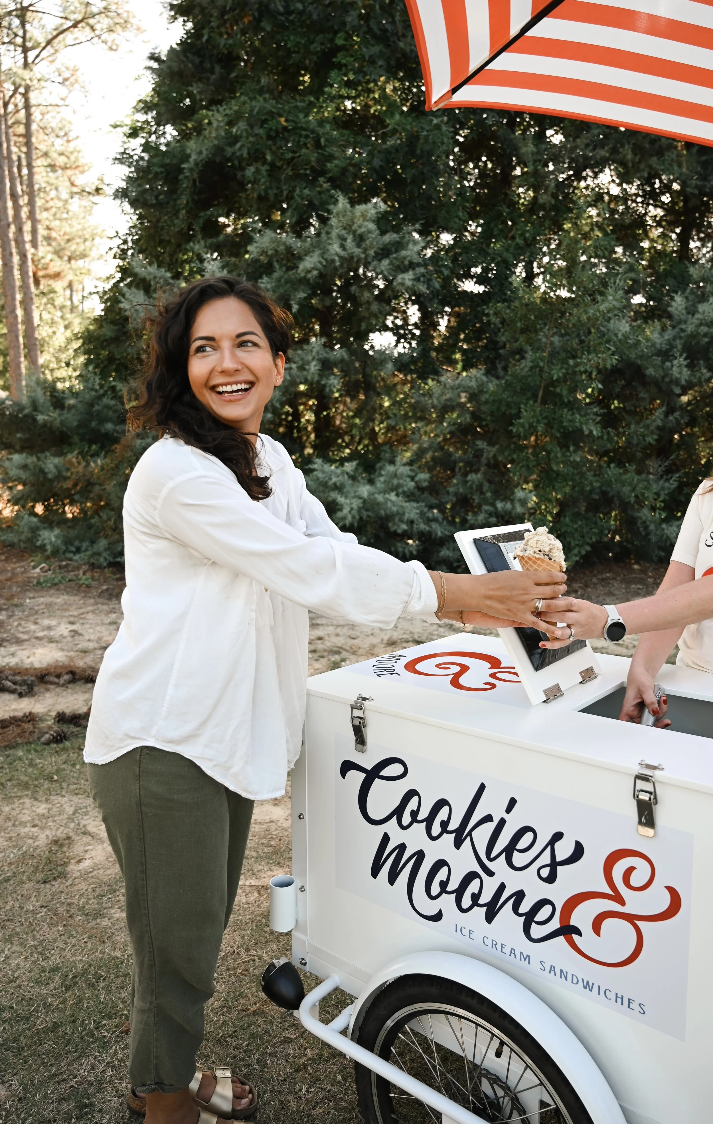 A woman in a white top at a private event with an ice cream bicycle labeled 'Cookies & Moore Ice Cream Sandwiches'. The background shows trees and an red-and-white striped umbrella. Rent the bicycle for your wedding today.