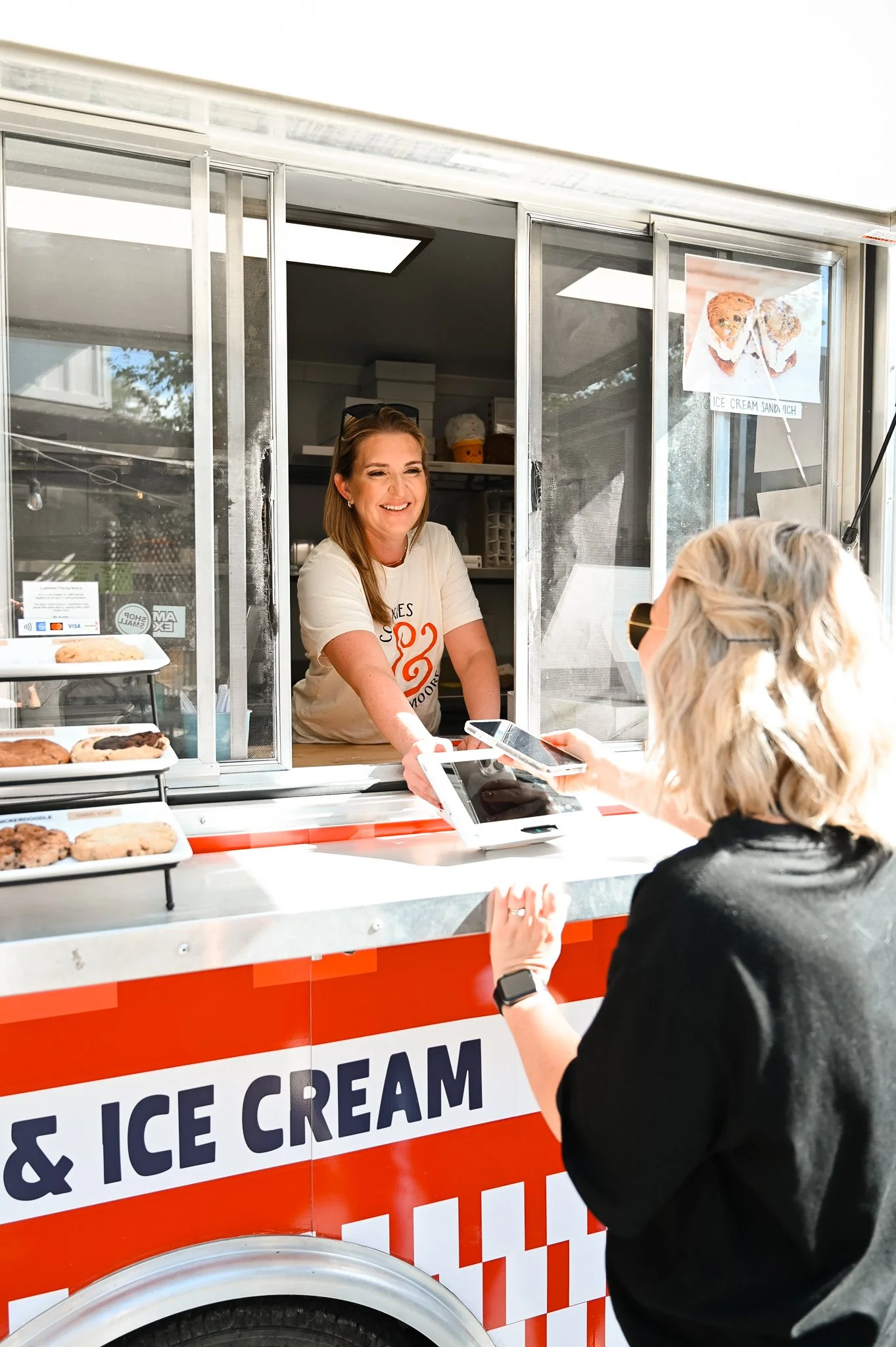 A woman at the Cookies & Moore Food Truck handing a receipt to a customer with blonde hair wearing sunglasses and a black top. The stand has a sign that says 'Cookies & Ice Cream' with a picture of cookies and ice cream sandwiches.