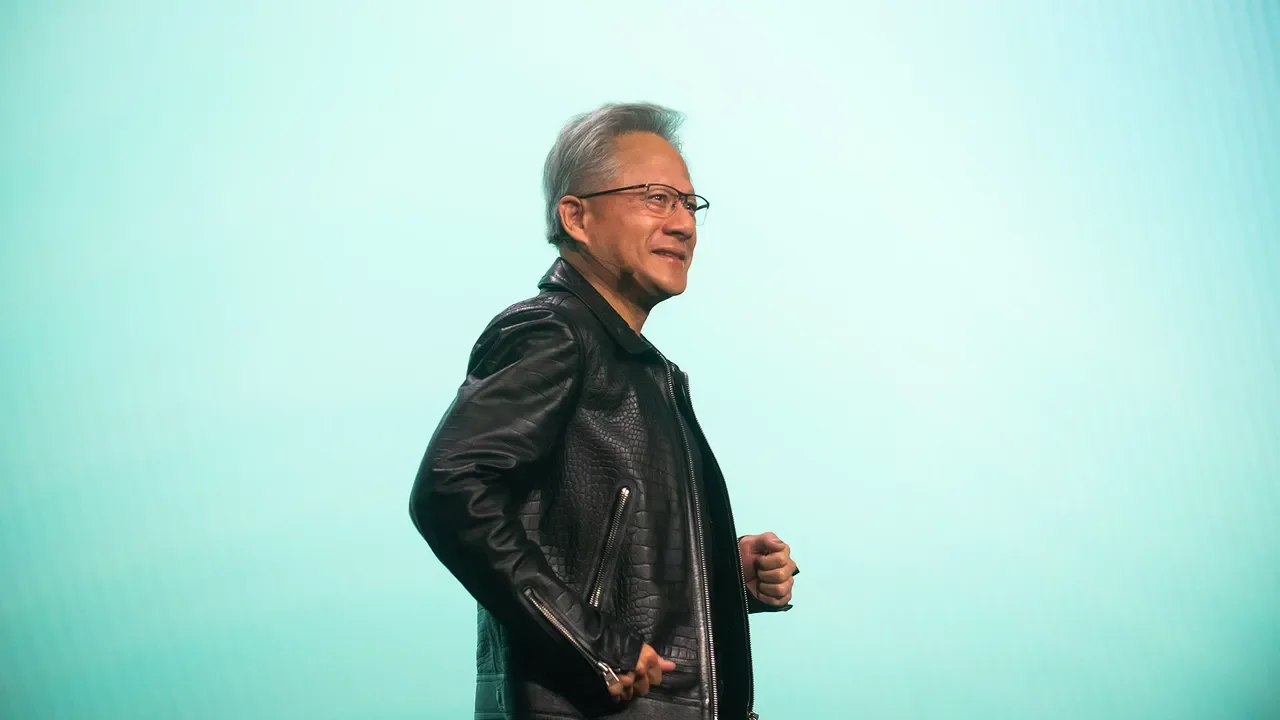 A middle-aged man with gray hair, wearing glasses and a black leather jacket, smiling and posing against a gradient teal background.