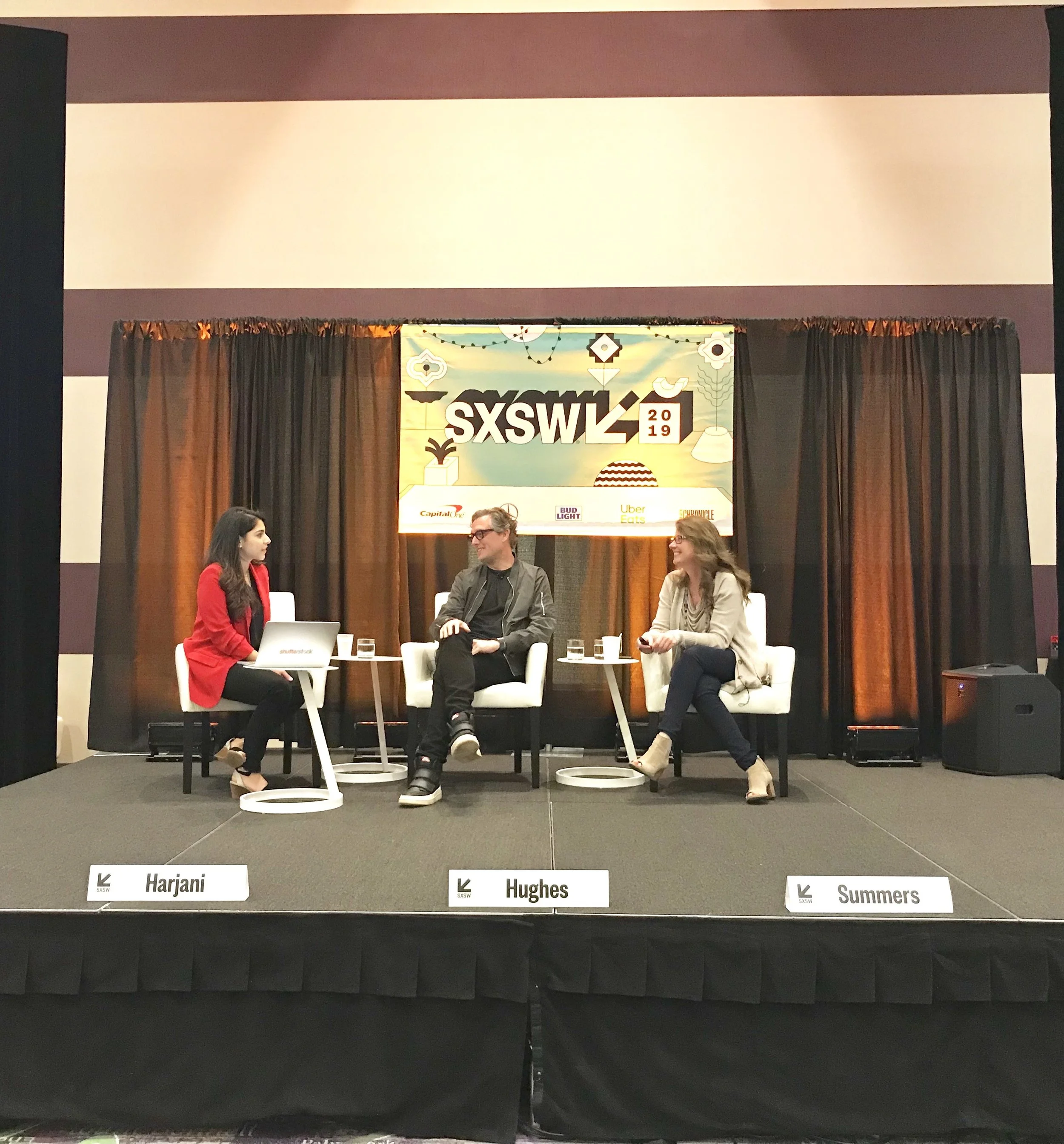 A panel discussion at SXSW 2019 with three people seated on stage. The stage has a backdrop with black curtains and a large SXSW 2019 sign. The panelists are identified as Harjani, Hughes, and Summers.