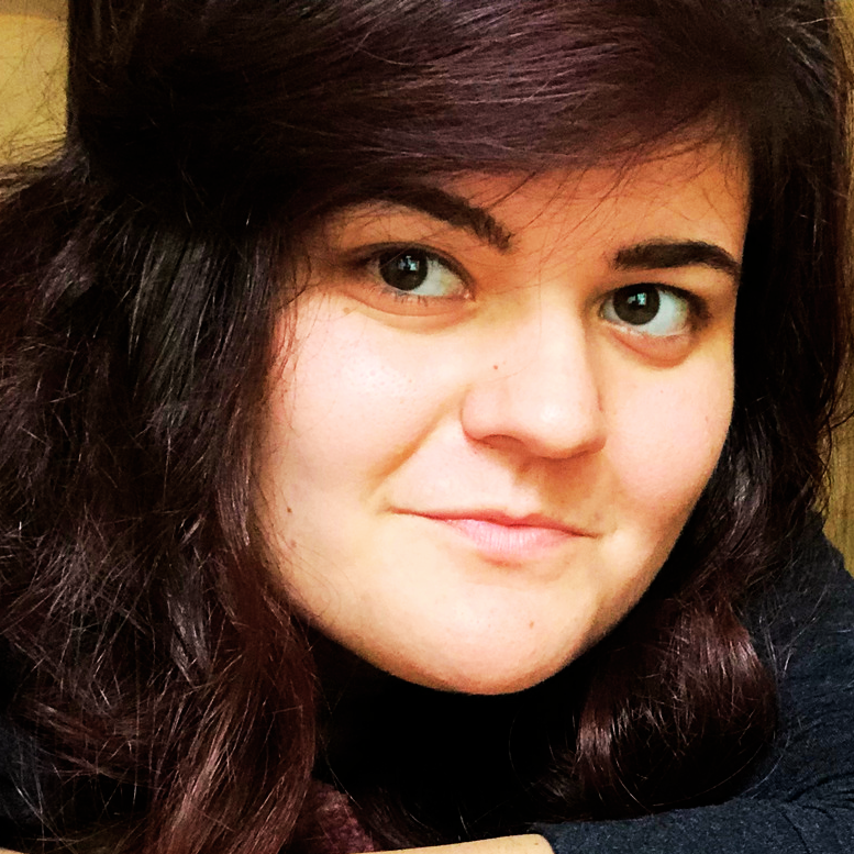 Close-up of a woman with dark brown curly hair, fair skin, and brown eyes, looking at the camera with a slight smile.