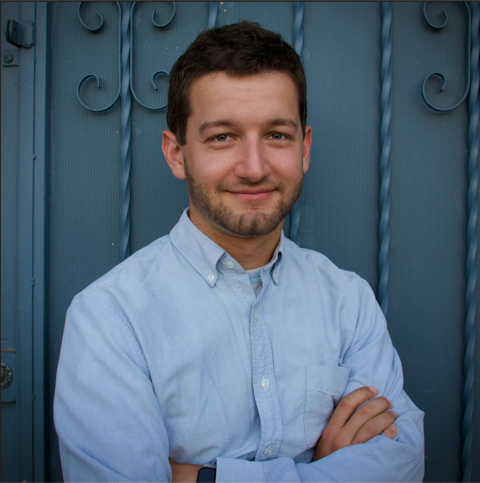 A young man with short dark hair and a beard, smiling with arms crossed, standing in front of a blue metal gate.