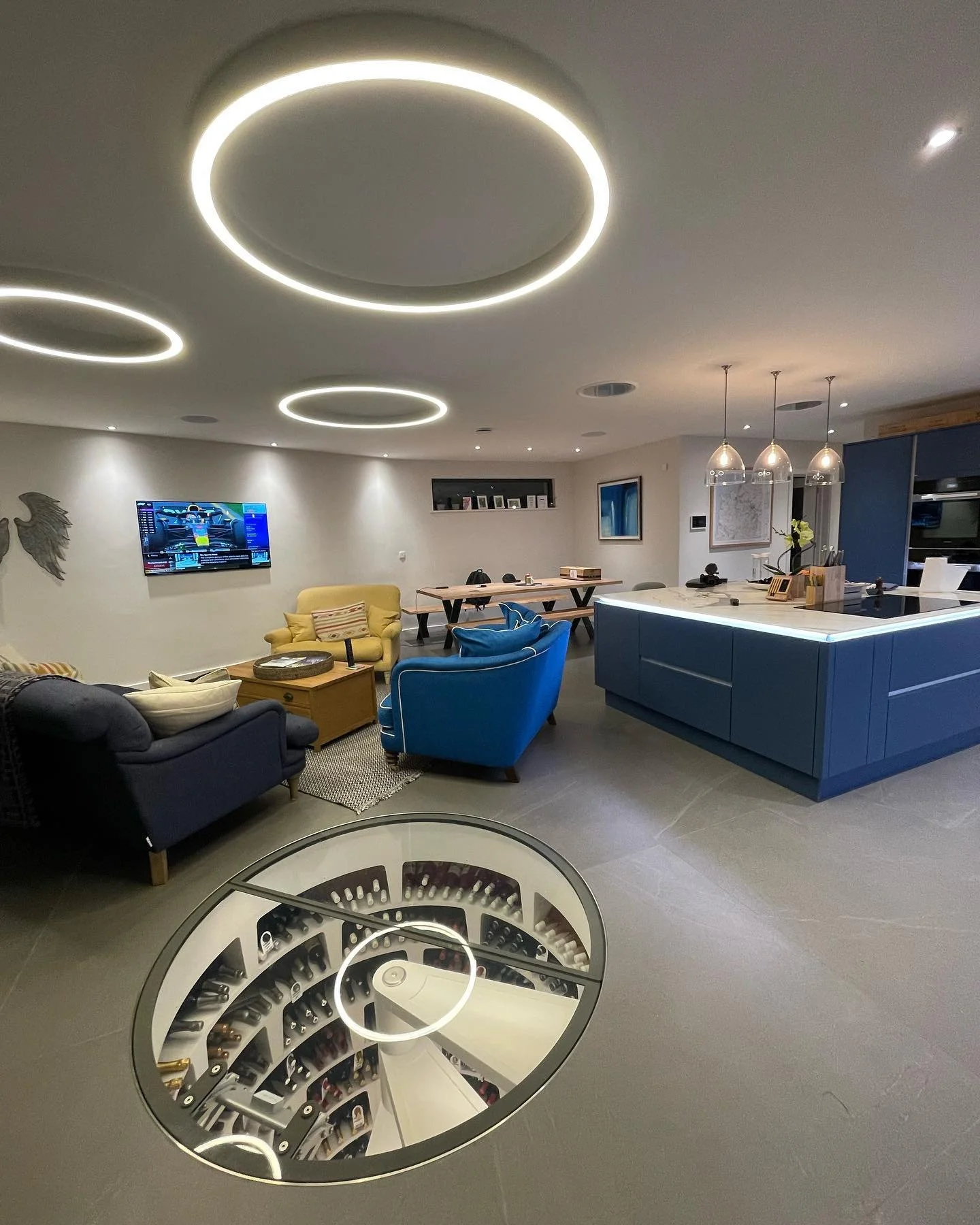 A modern living space and kitchen with circular ceiling lights, a wall-mounted TV, blue and yellow armchairs, a chandelier, and a glass floor window revealing a wine cellar underneath.