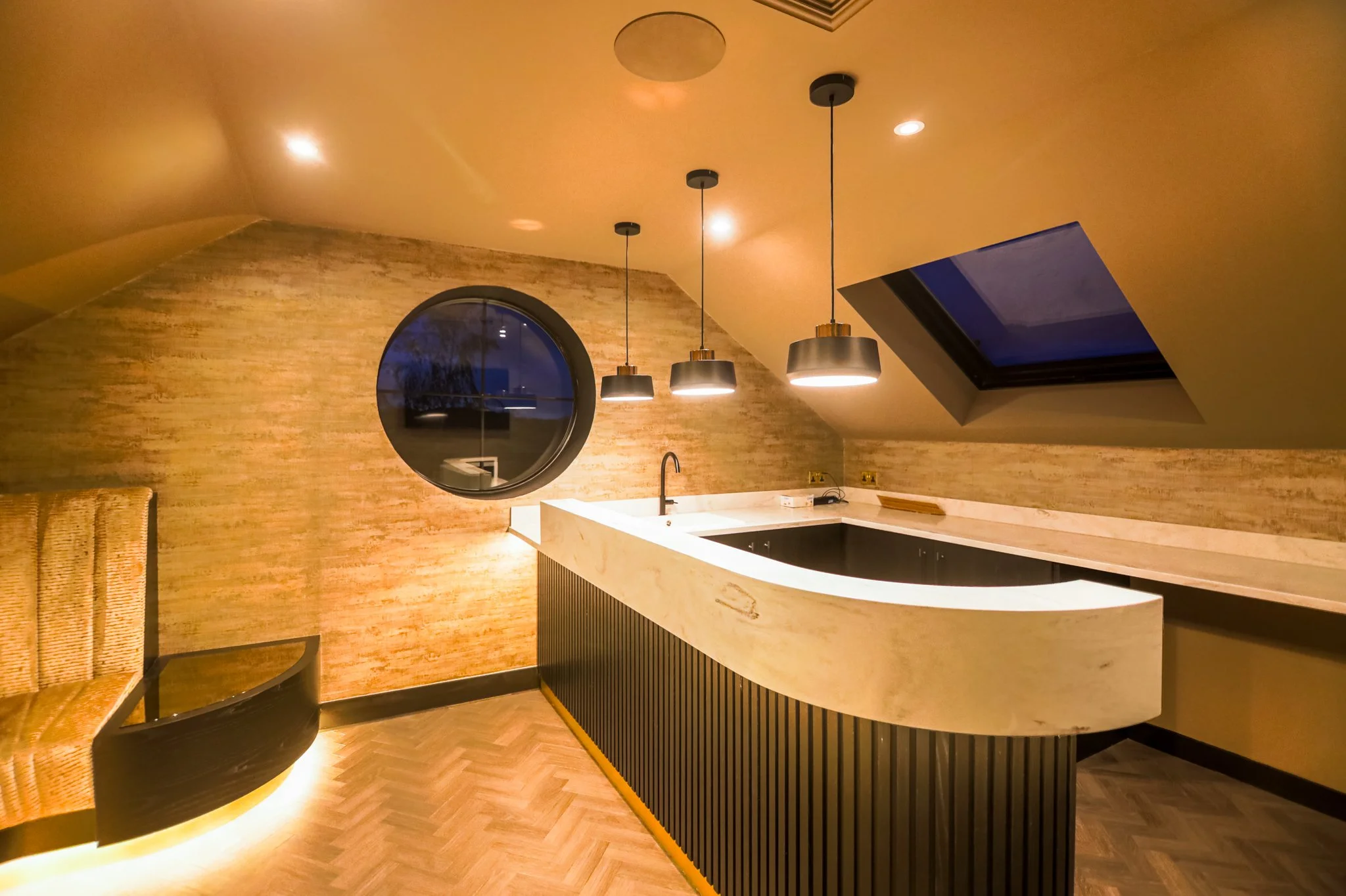 A modern attic kitchen with a sloped ceiling, circular window, skylight, hanging pendant lights, marble countertops, black backsplash, and a wooden chair.