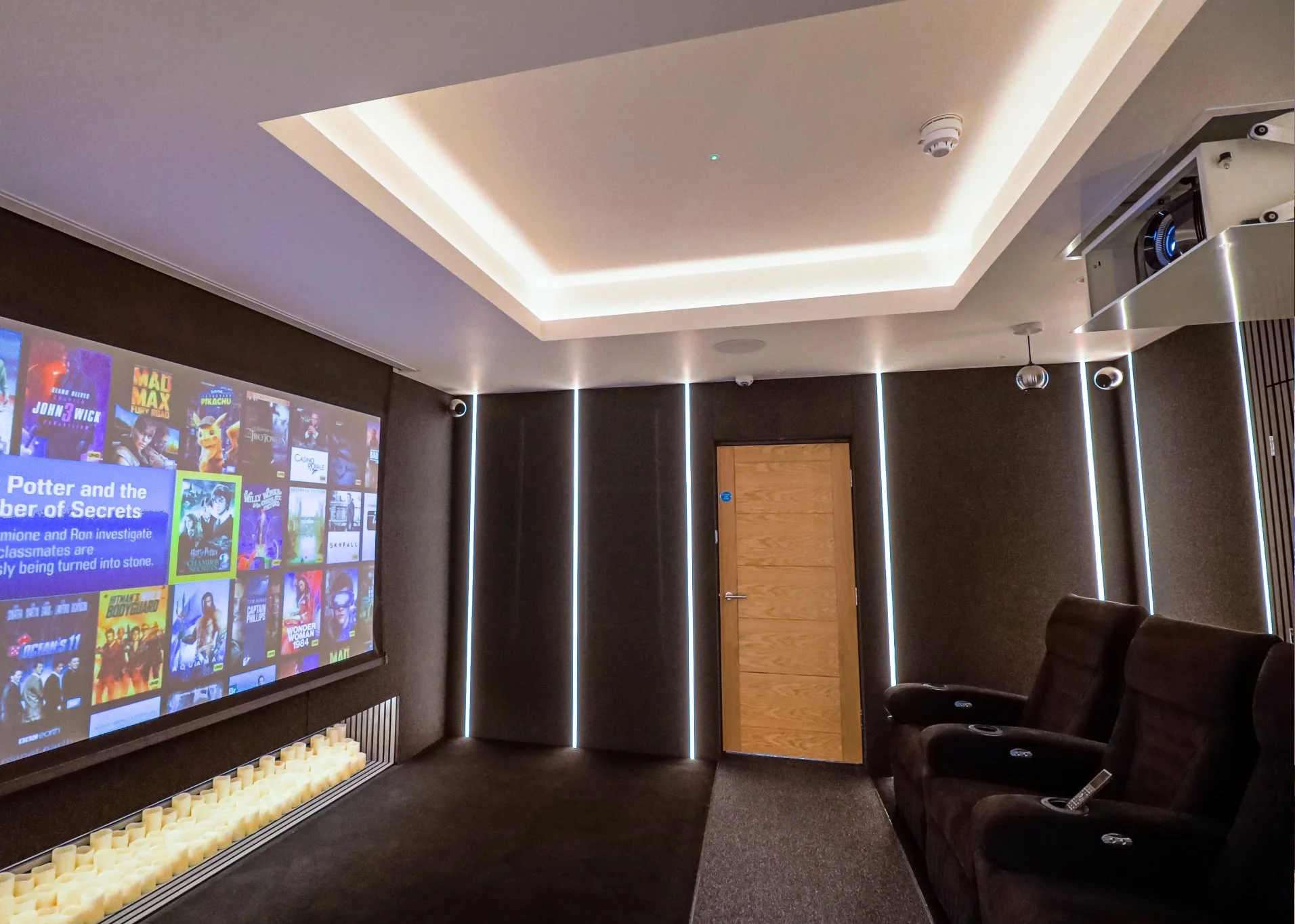 A home theater room with a large screen showing movie posters, dark walls with vertical white light accents, a wooden door, a ceiling with recessed lighting, and three brown reclining chairs.