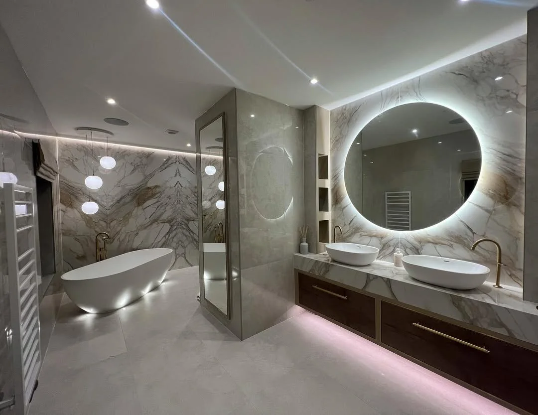 Modern bathroom with marble walls and floors, featuring a freestanding bathtub, double vanity with vessel sinks, large backlit mirror, glass shower enclosure, and decorative lighting.