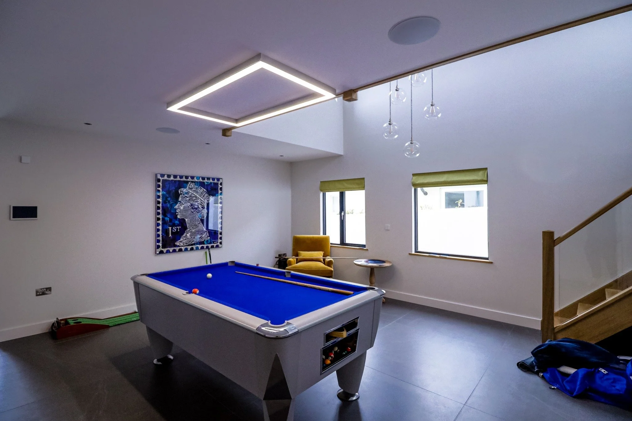 Modern game room with a pool table, a yellow armchair, and two windows with green blinds.