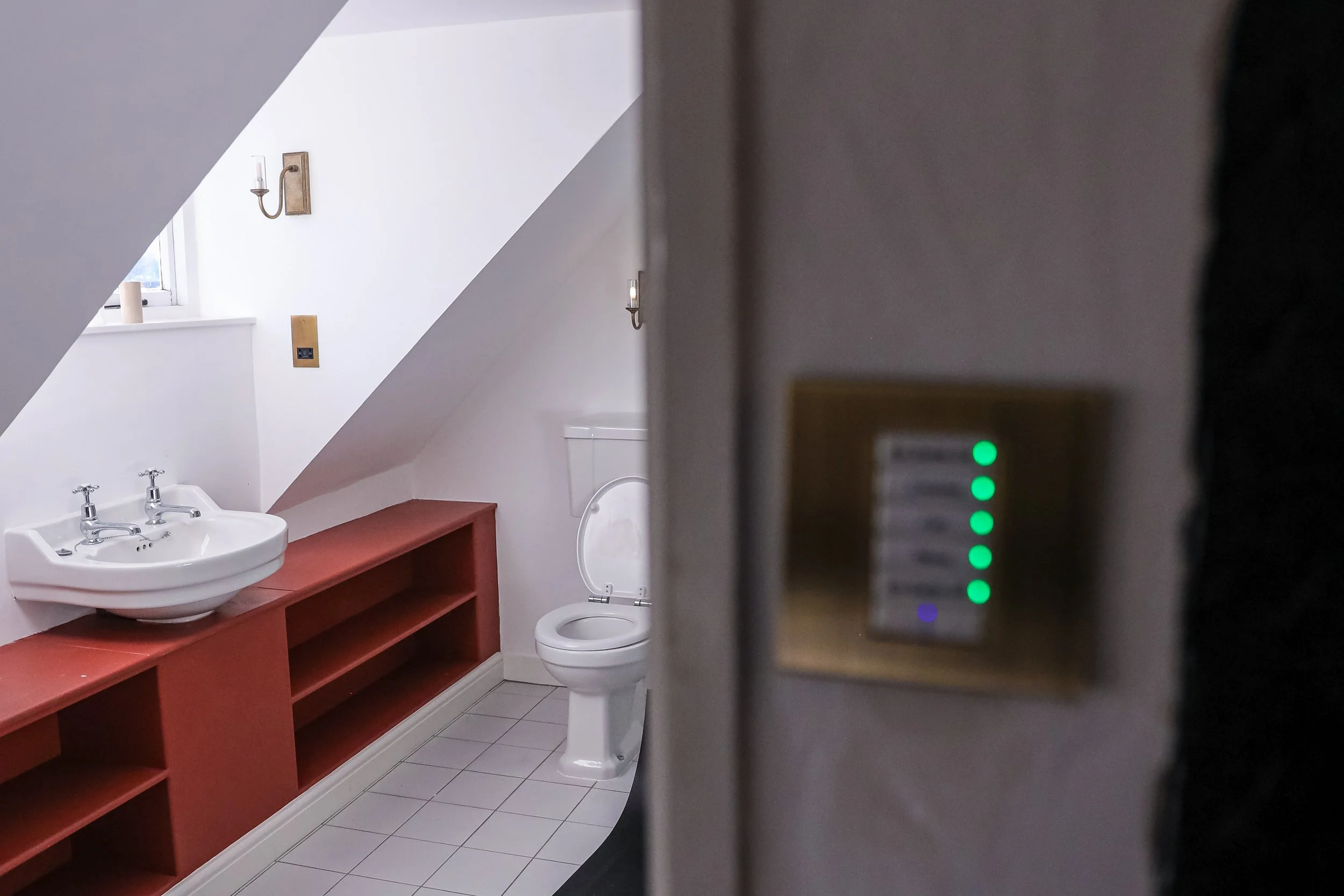 Bathroom with sink, toilet, wooden storage, sloped ceiling, and wall-mounted lights; viewed through a partially open door with indicator lights.